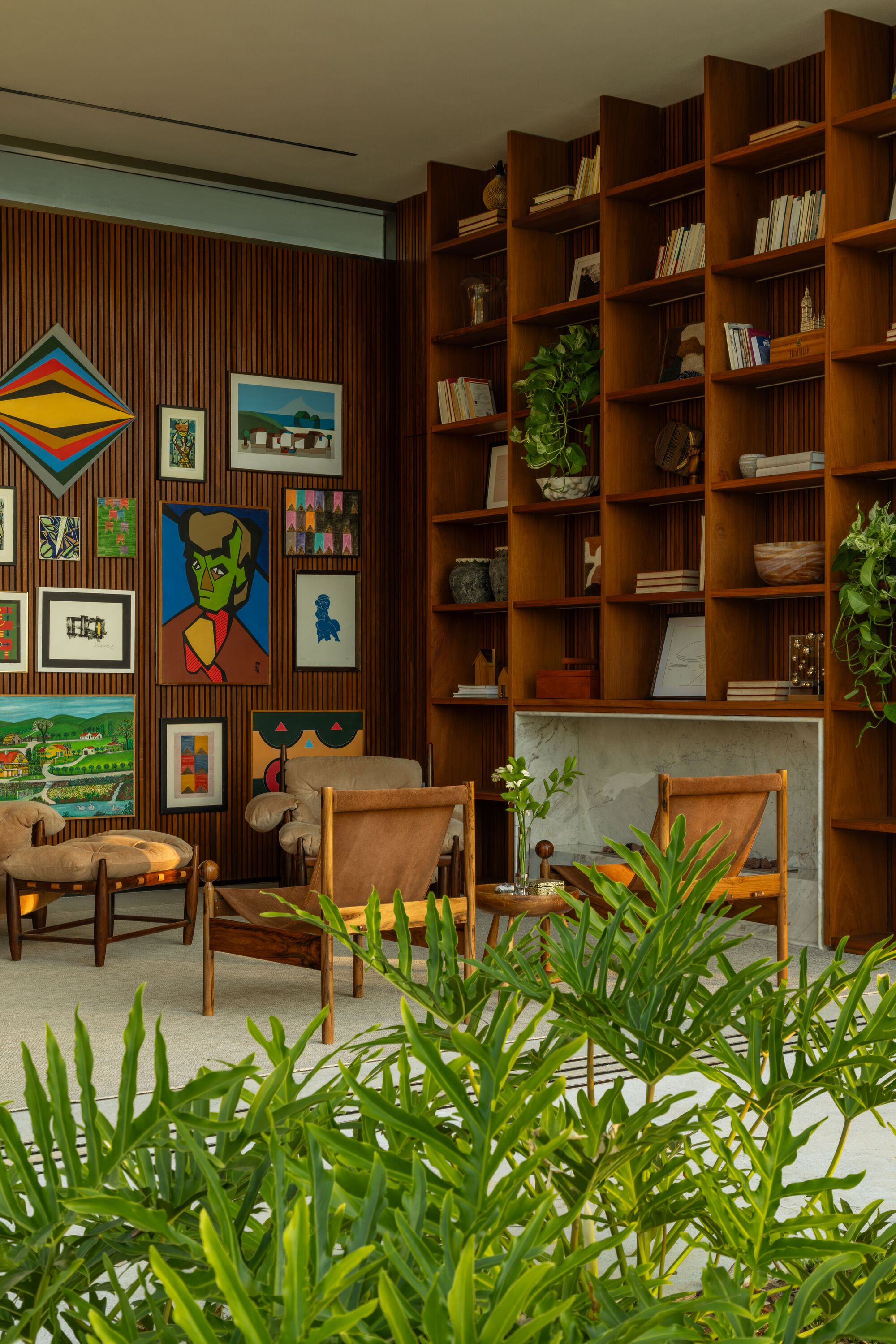 Timber bookshelf wall beside a gallery of framed artworks with potted palms in foreground