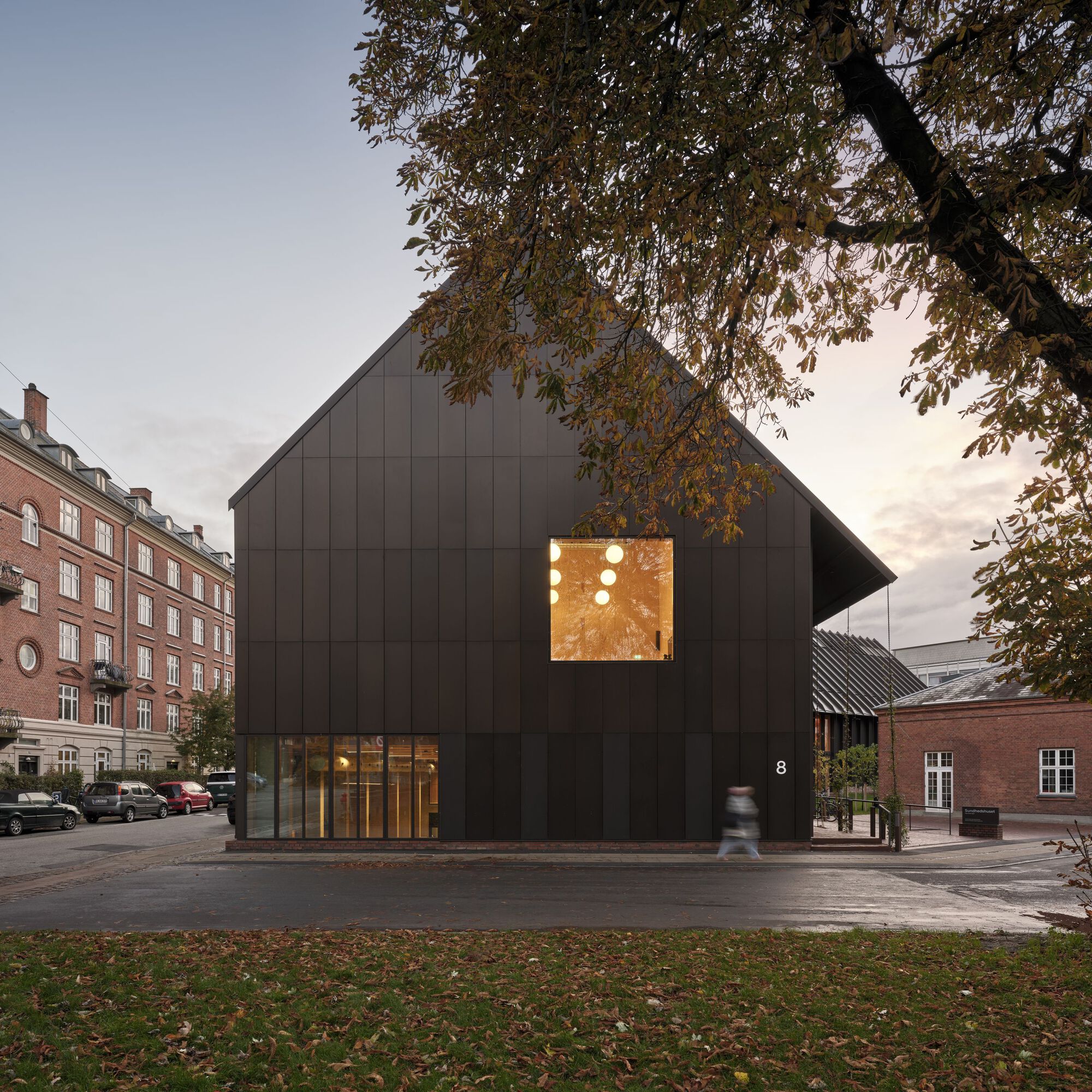 Dark metal-clad gabled facade with illuminated upper window and ground-level glazing at dusk