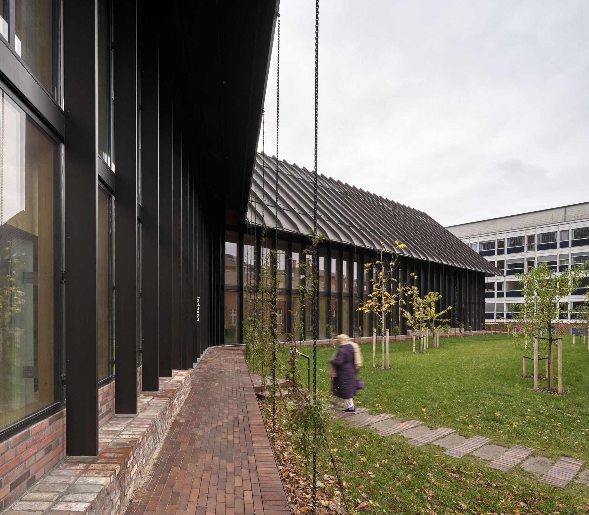 Brick walkway along black metal facade with young trees and a person passing by
