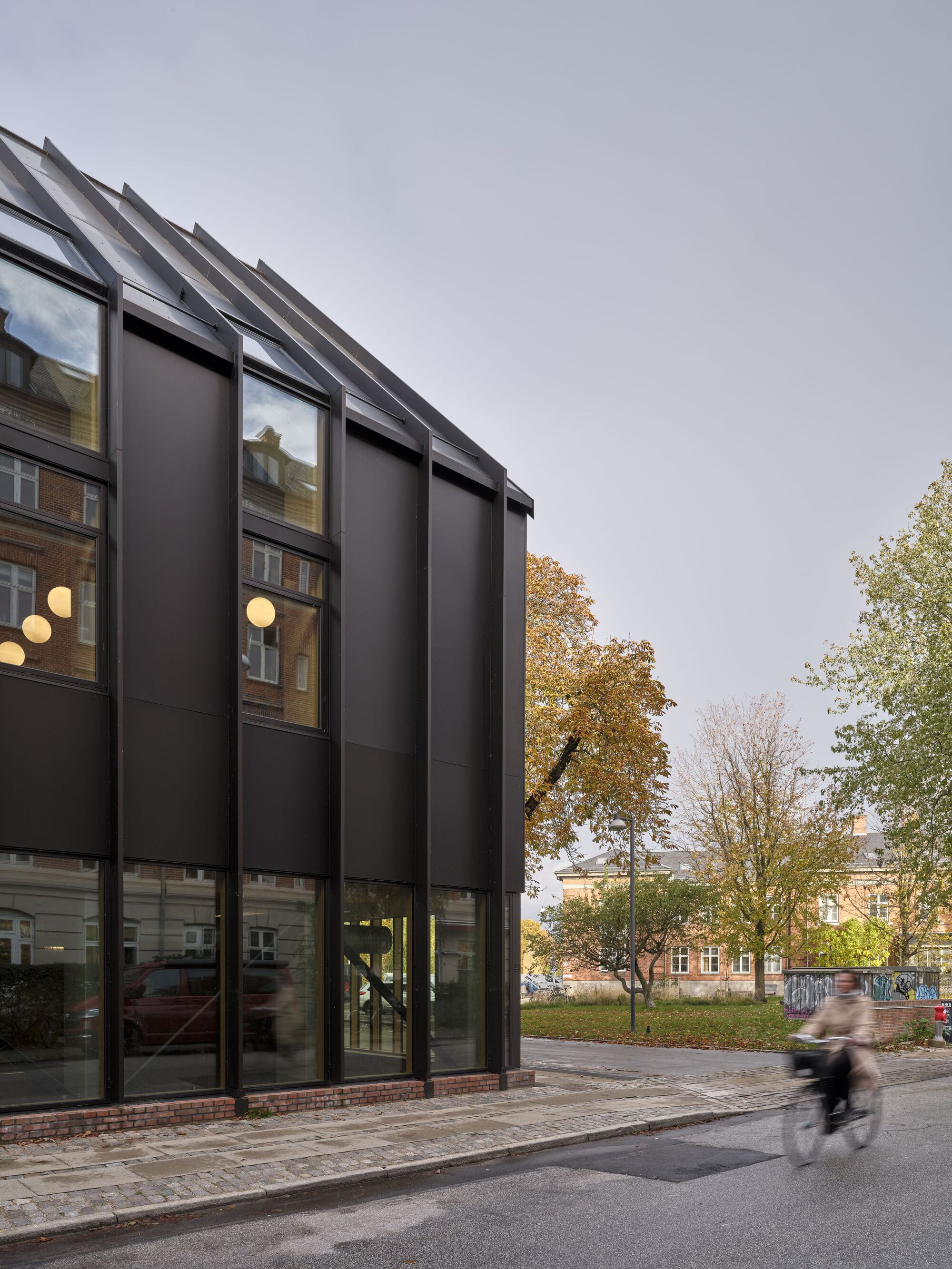 Street view of the black metal panel facade with vertical glazing and a cyclist passing in motion blur