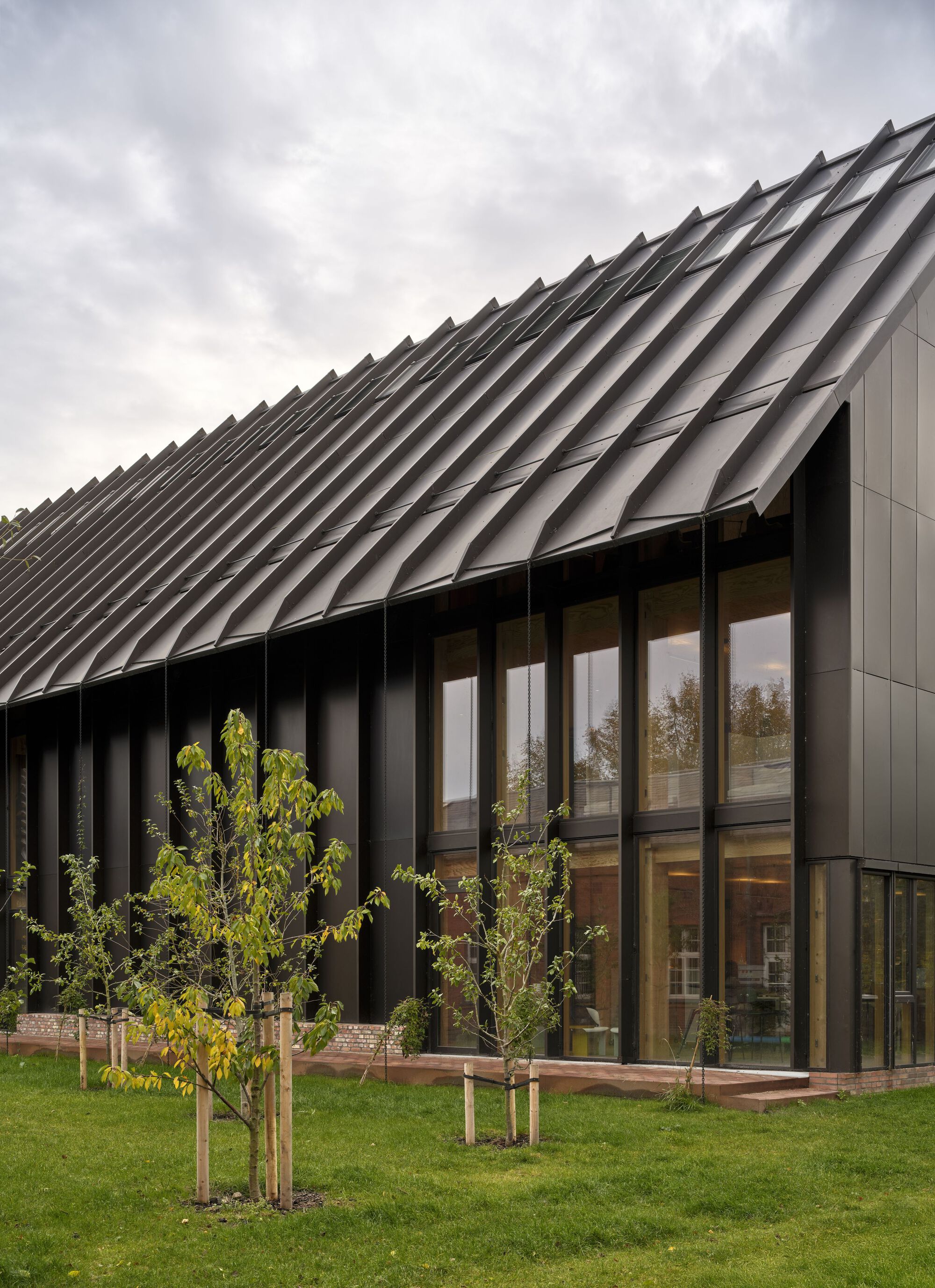 Sawtooth metal roof above glazed facade and black vertical fins with young trees on lawn in autumn