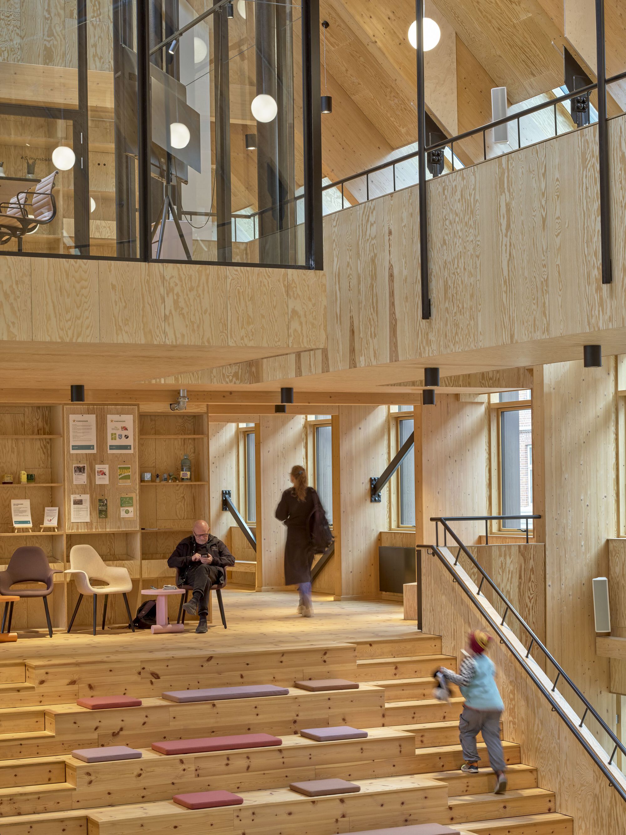 Tiered seating area with plywood benches and visitors moving through the double-height plywood-lined space