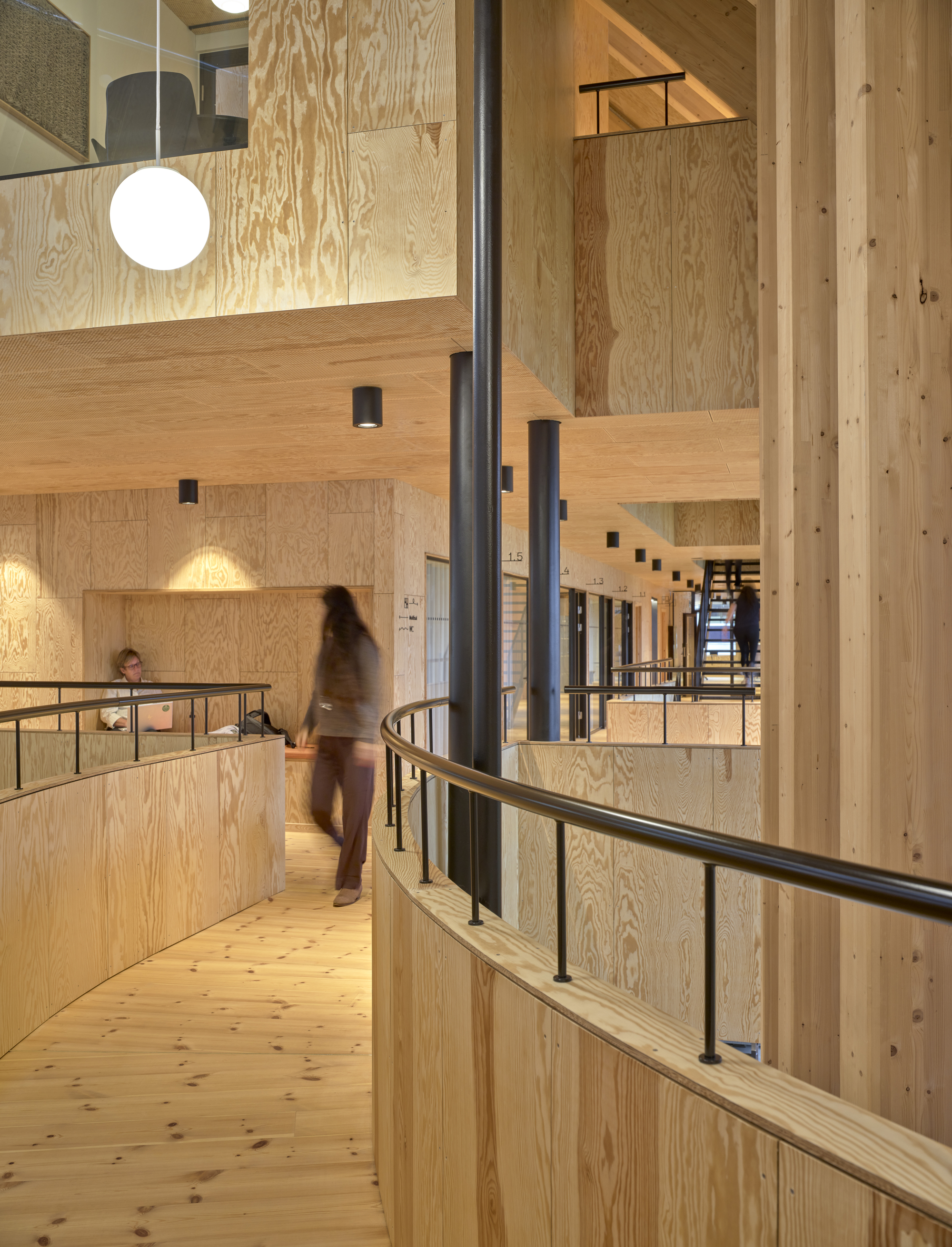 Curved balcony with black steel railings and a person in motion crossing the plywood-lined circulation level