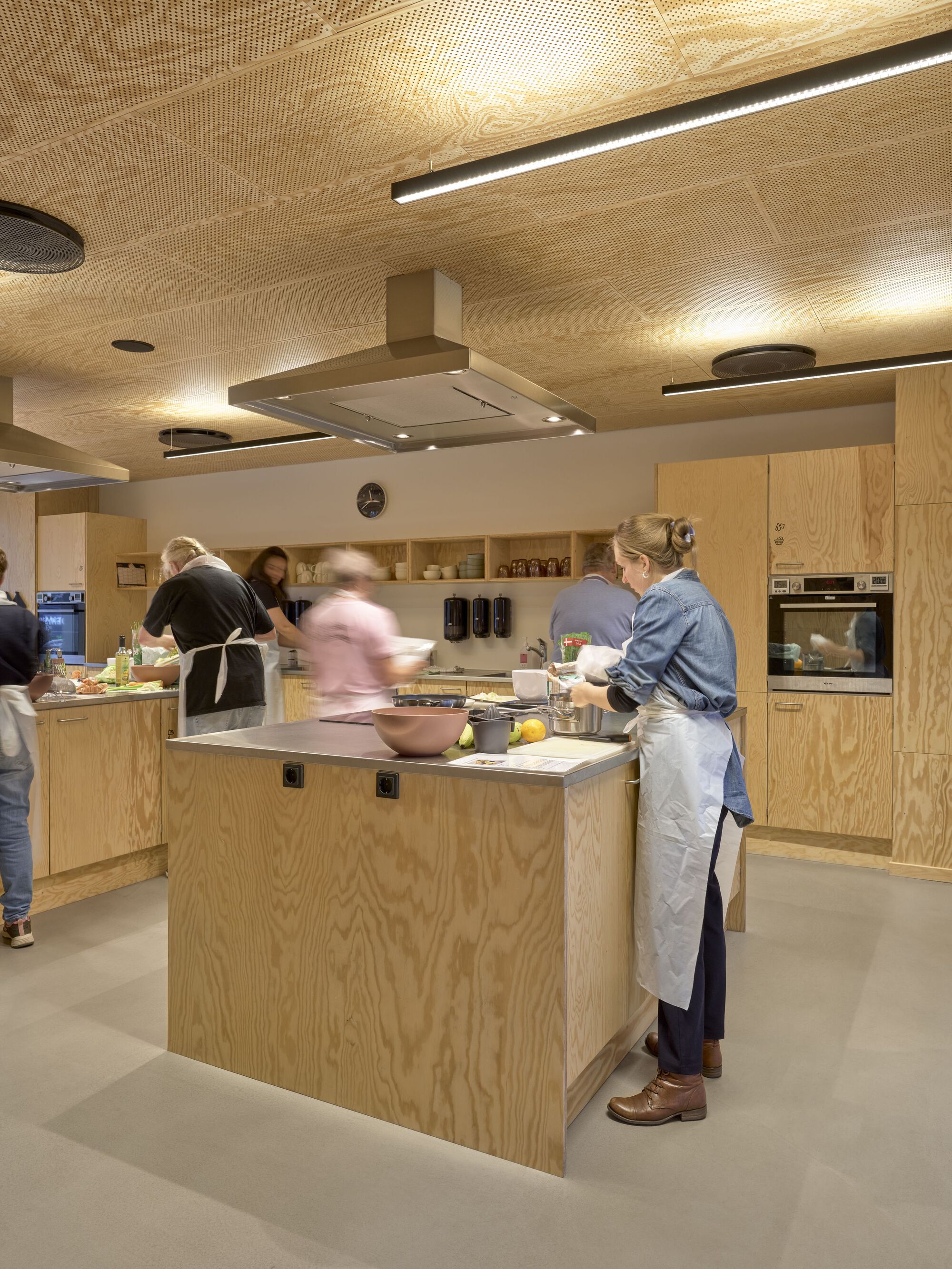 Communal kitchen with plywood island and cabinetry as people prepare food beneath a perforated timber ceiling