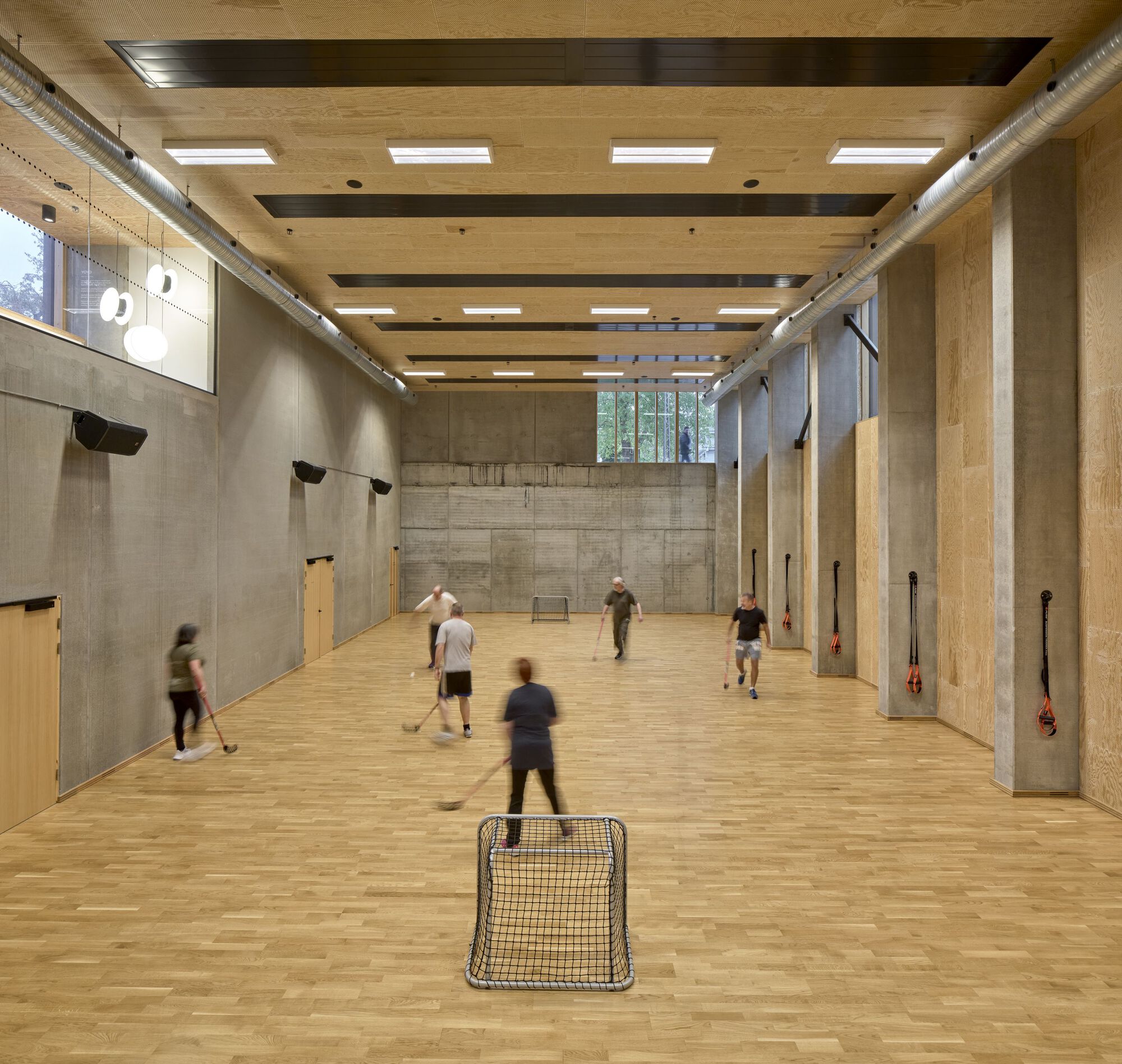 Indoor sports hall with board-marked concrete walls, timber ceiling beams and players in motion blur