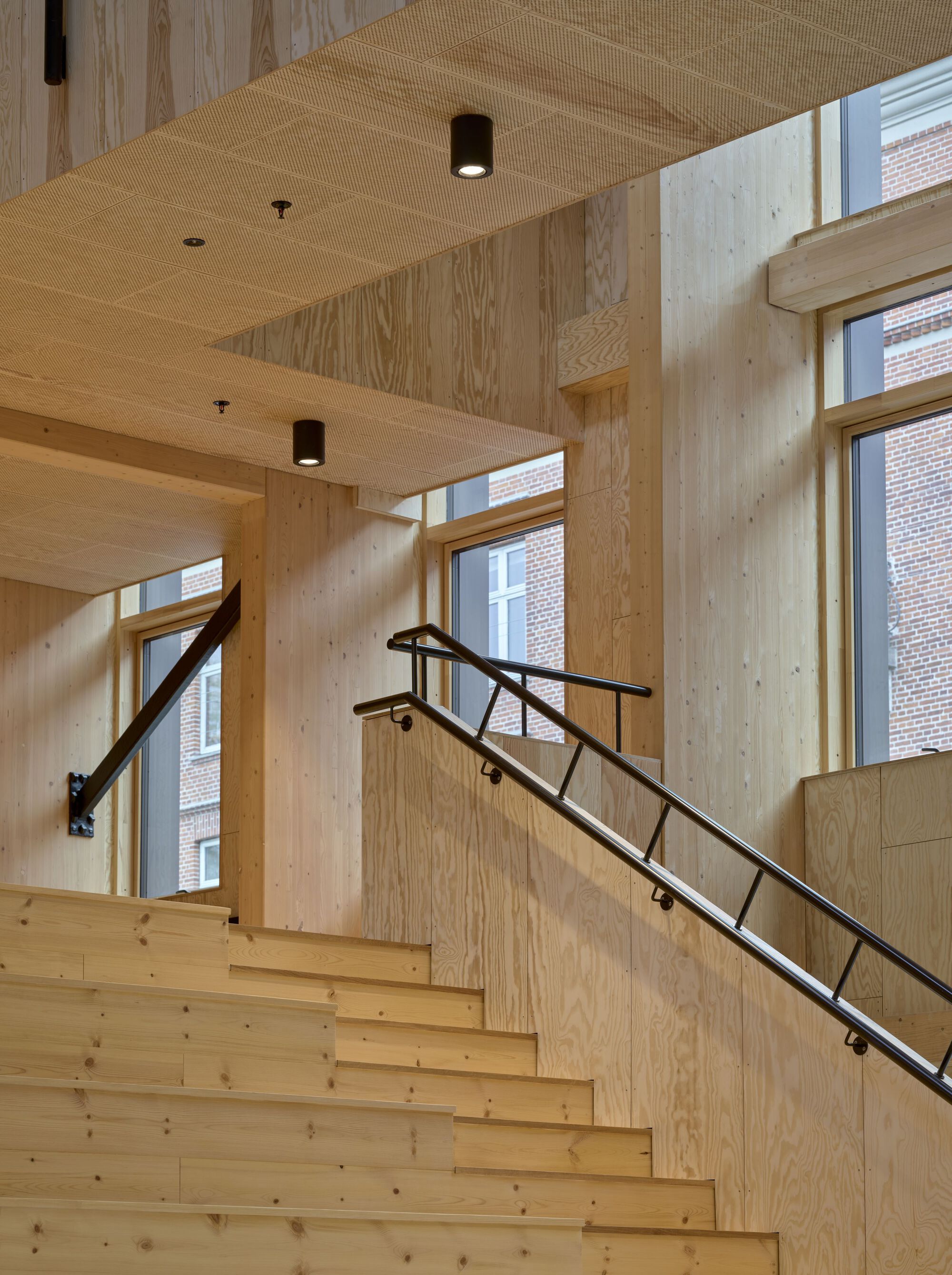 Wood-clad staircase with black metal railing bathed in afternoon sunlight from tall windows