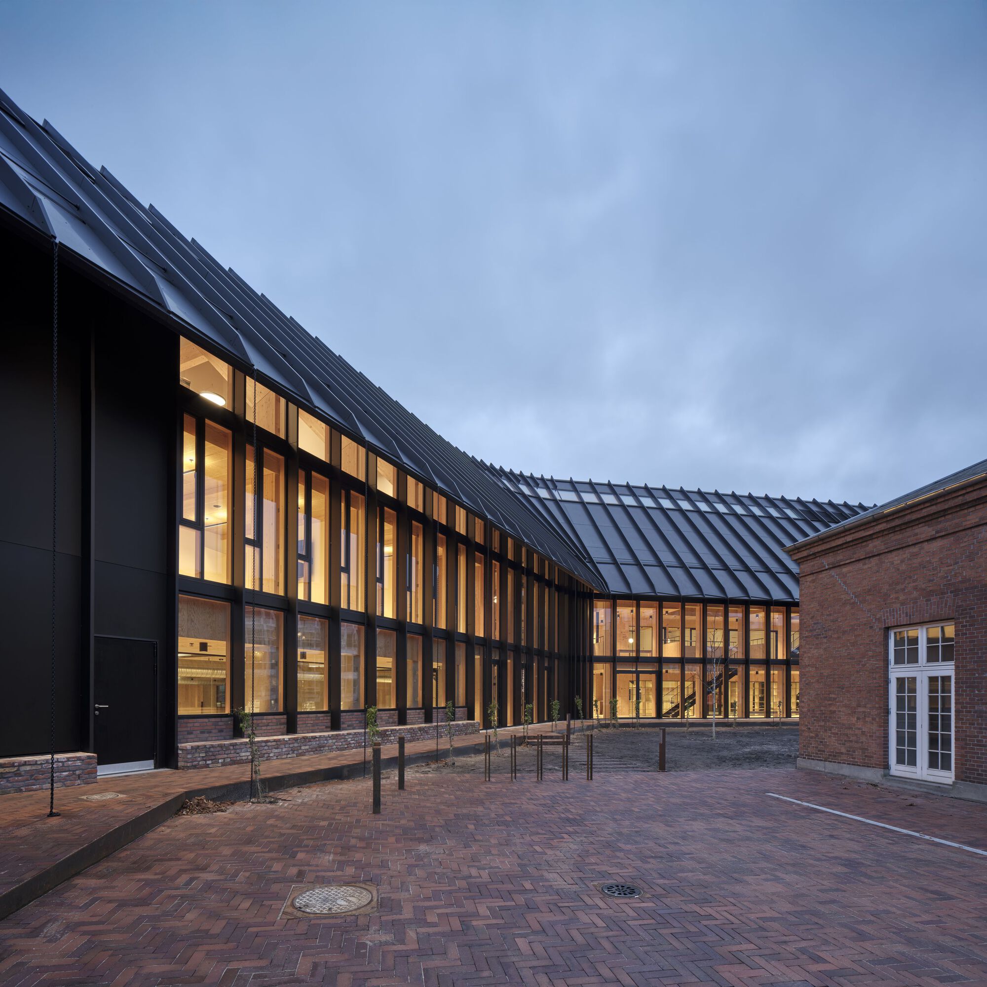 Brick plaza at dusk framed by the angular metal roof and warmly lit interiors