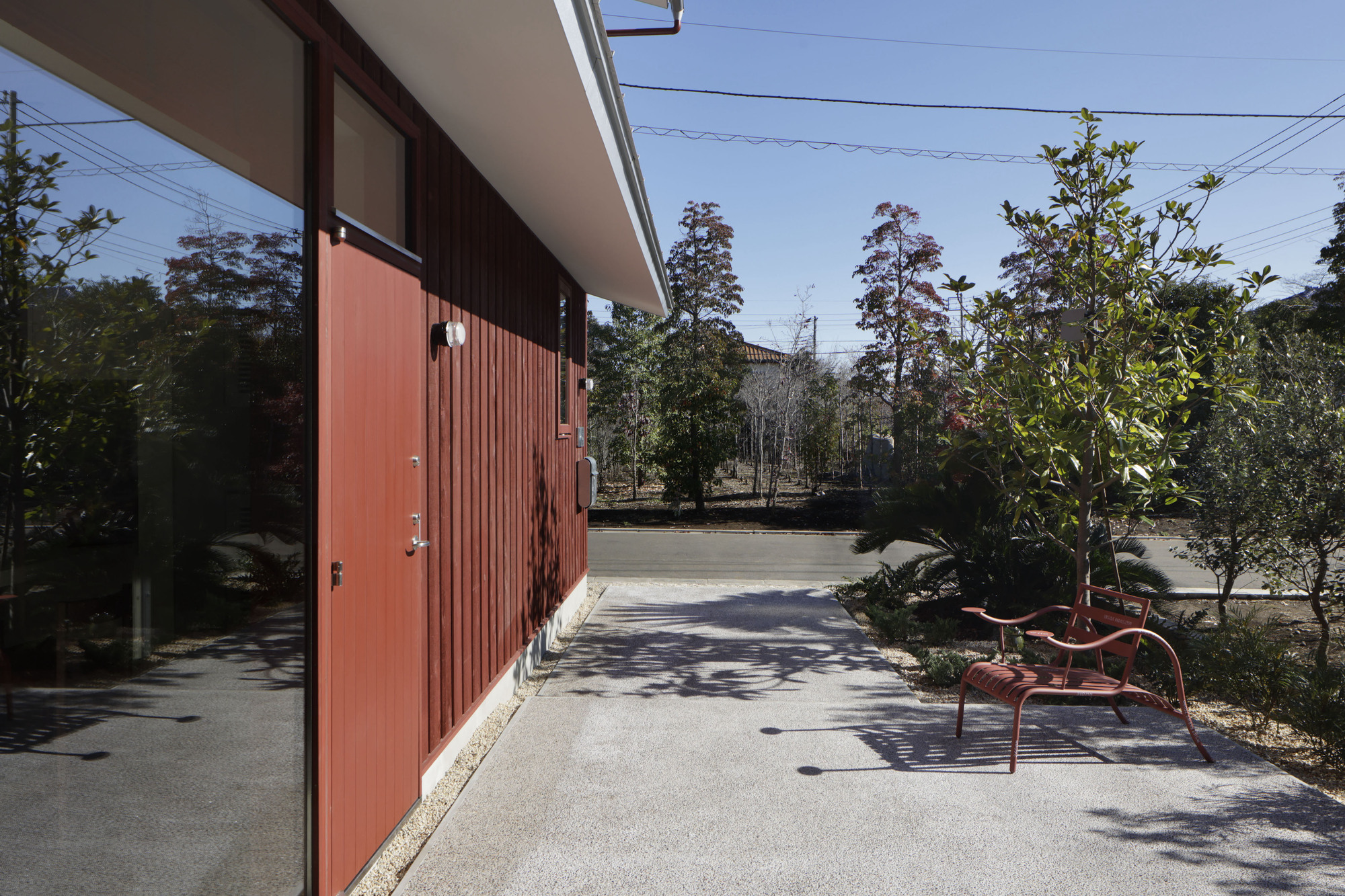 Courtyard with vertical timber cladding, concrete paving, planted trees and red bench in dappled shade