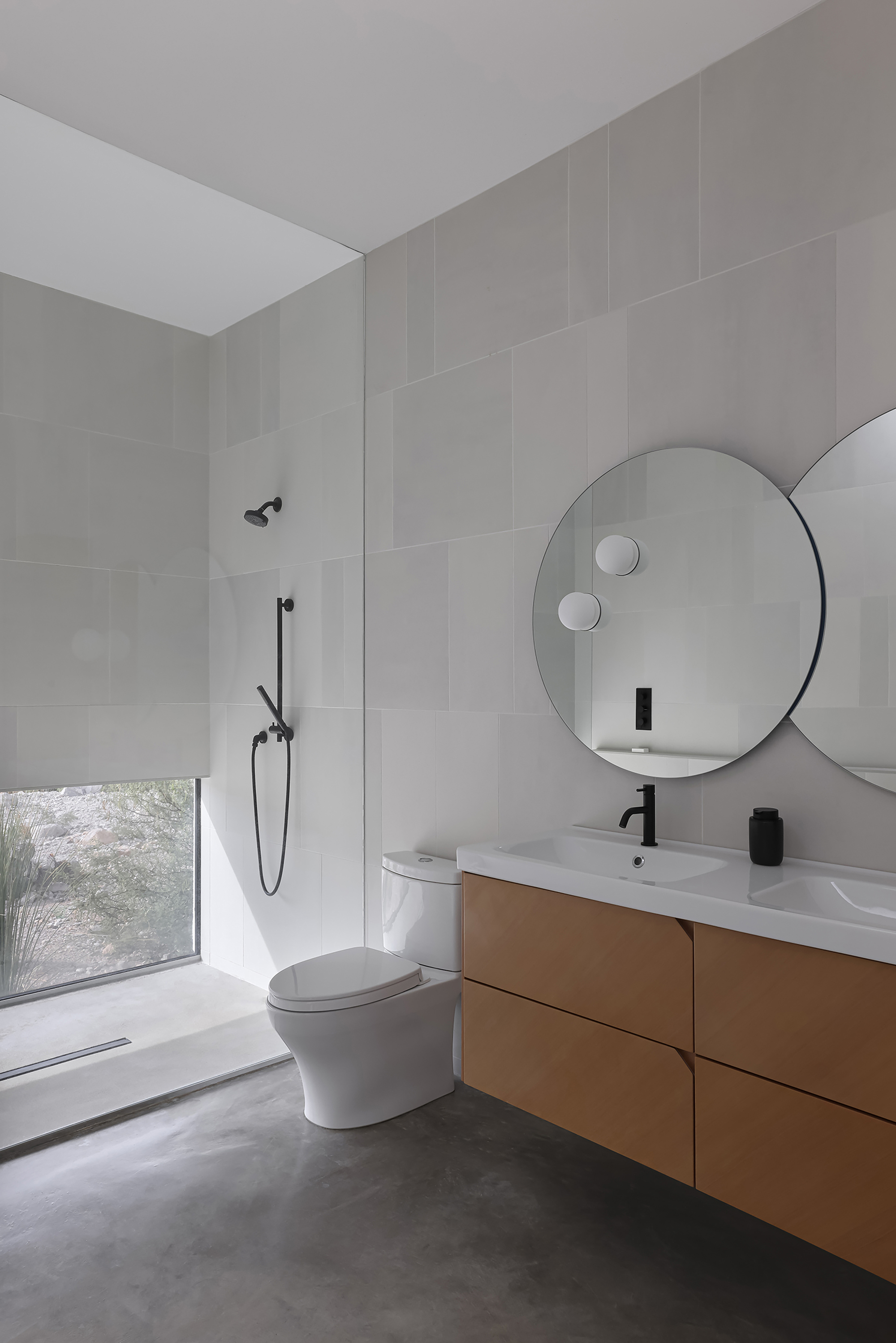 Bathroom with circular mirror above timber vanity and low desert window with roller shade