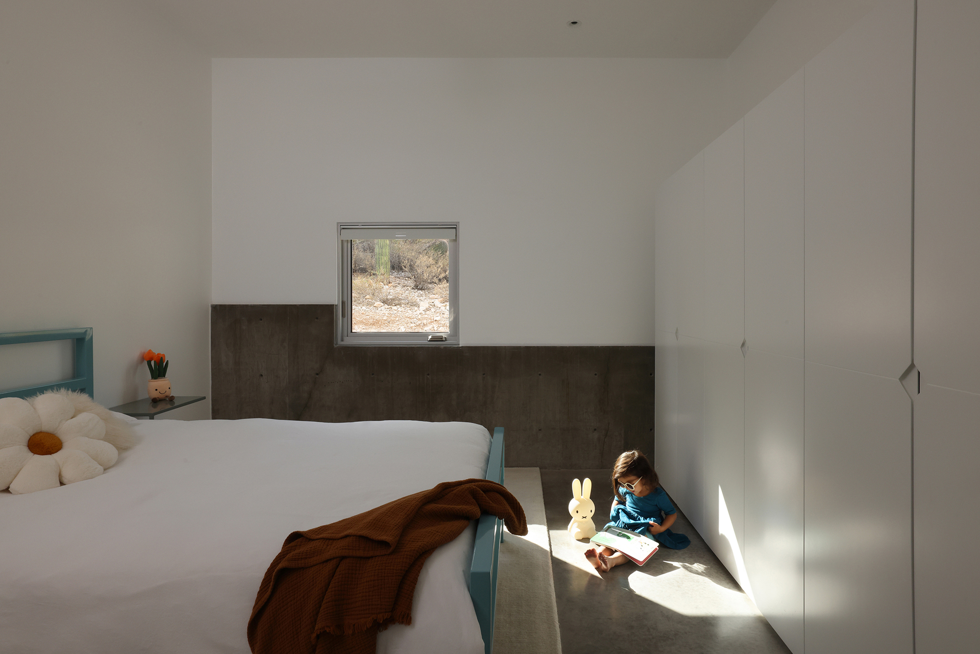 Child seated in sunlit corner beside bed with metal wainscoting and square desert window