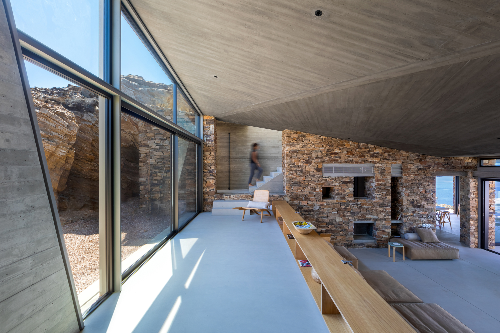 Interior living space with exposed stone walls, concrete ceiling, and glazed corner framing a figure in motion
