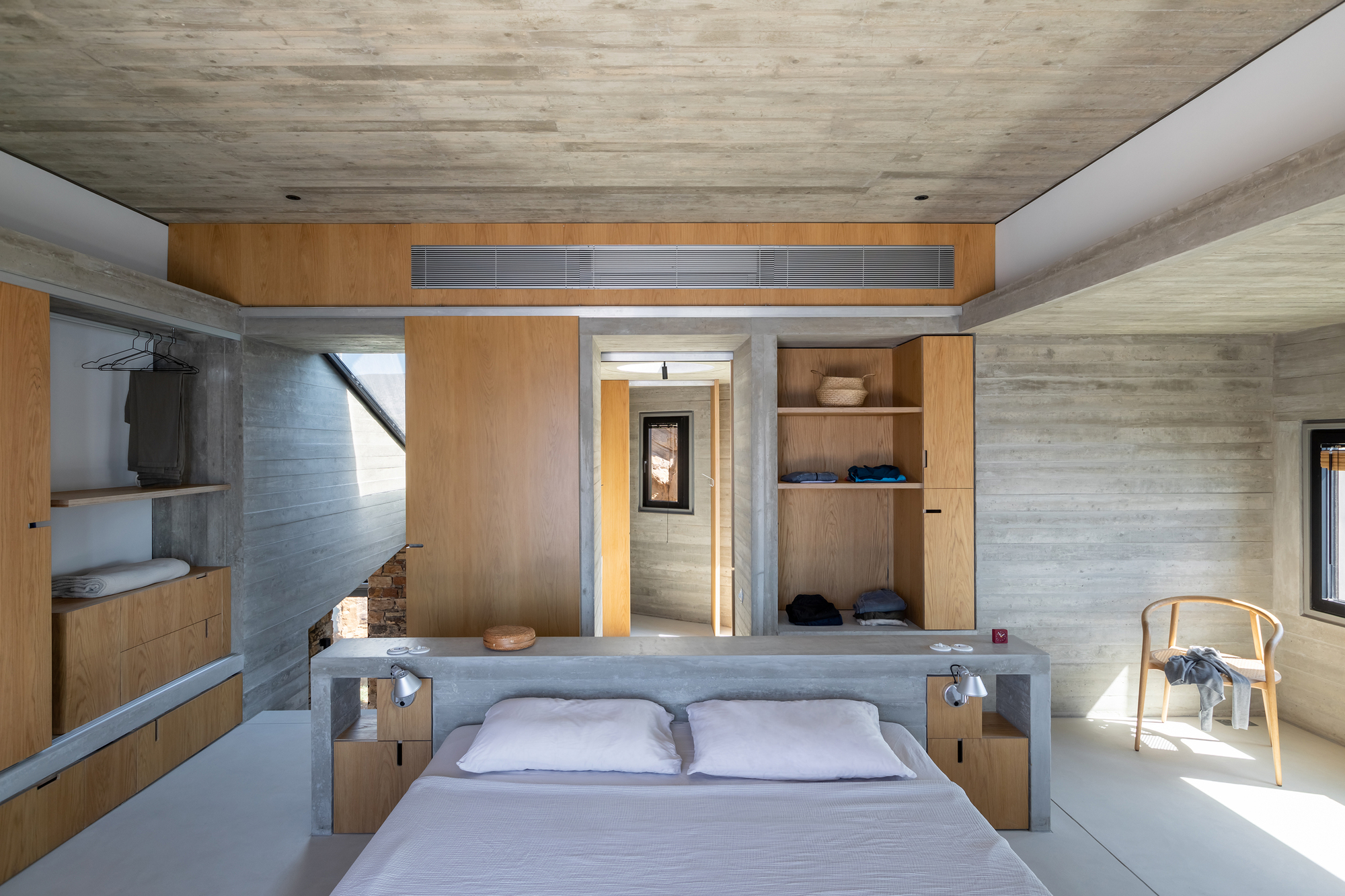 Bedroom with board-formed concrete walls, timber built-in wardrobes, and a doorway framing the passage beyond