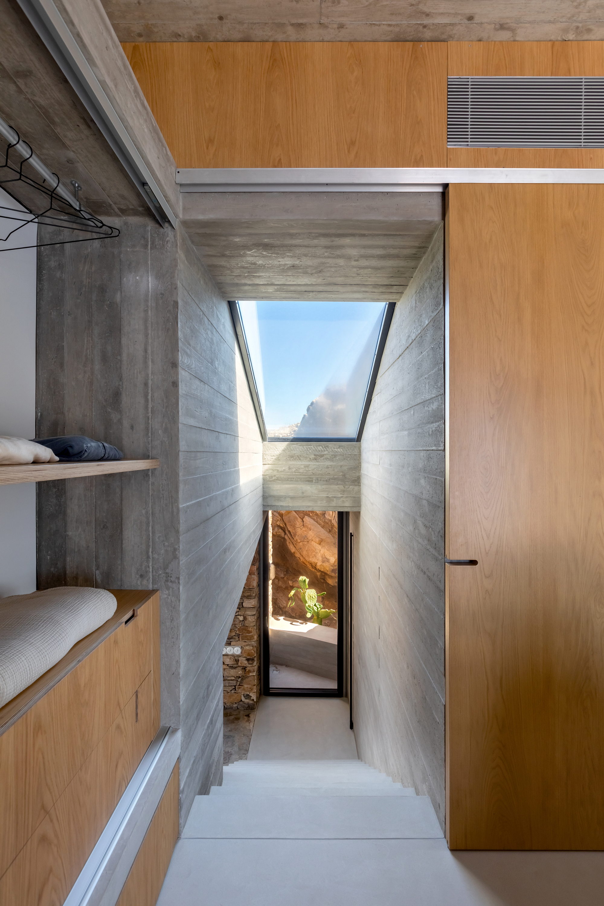 Corridor with angled skylight above board-formed concrete walls leading to a doorway with rock outcrop visible