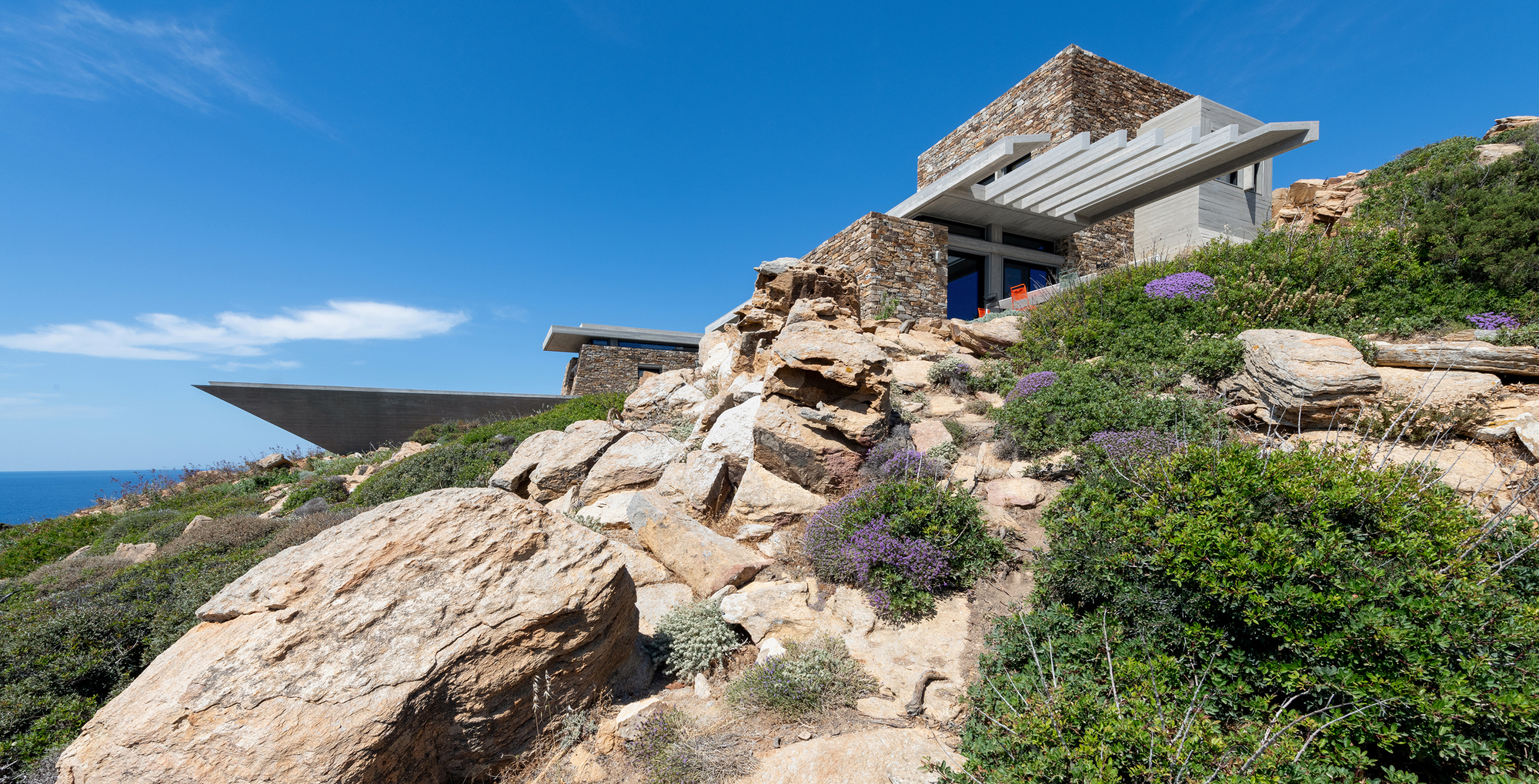 Stone-clad volumes terraced into the hillside with native vegetation and boulders under a clear sky