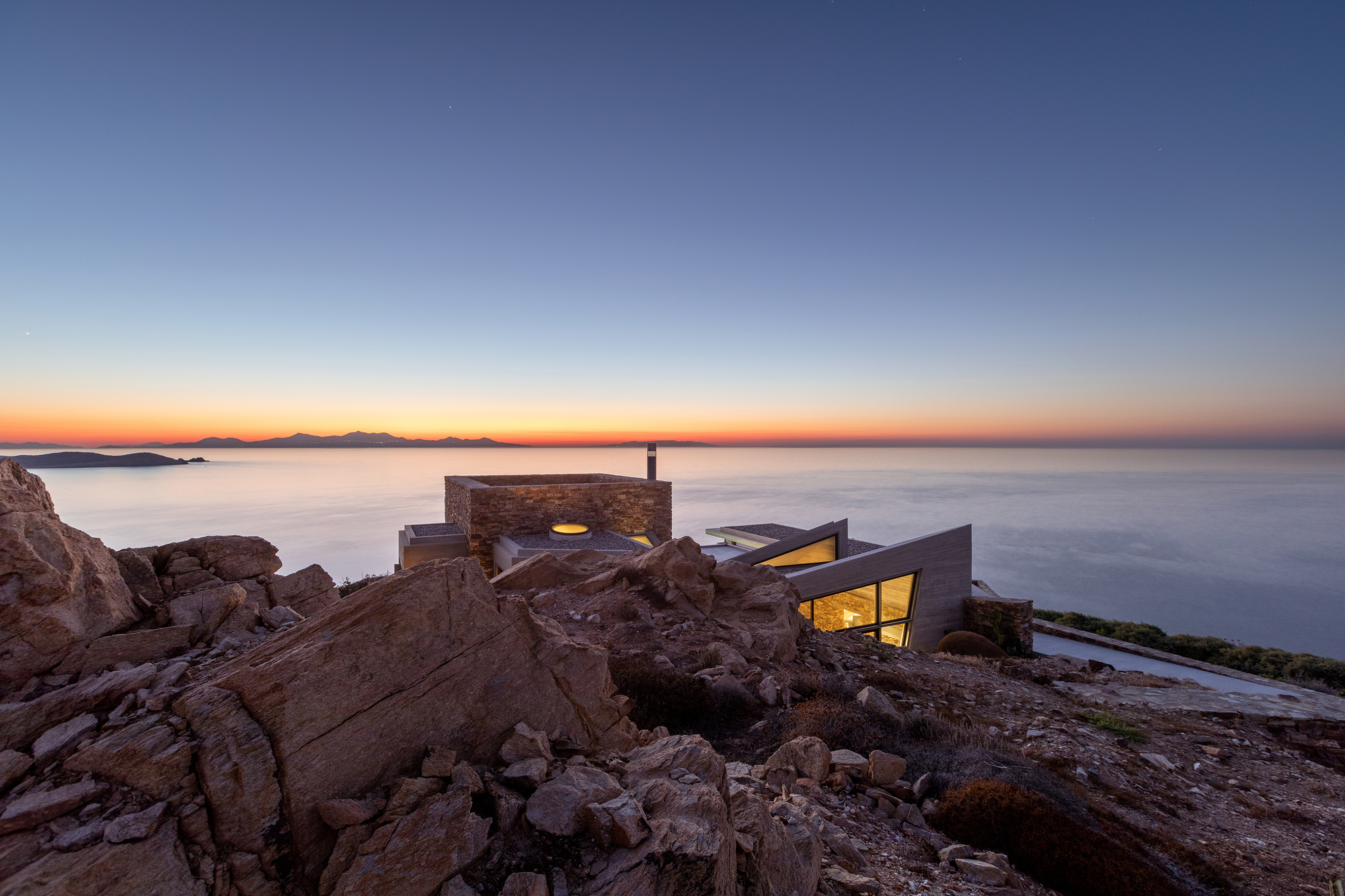 Stone volumes with illuminated windows perched on rocky outcrops at dusk above the water