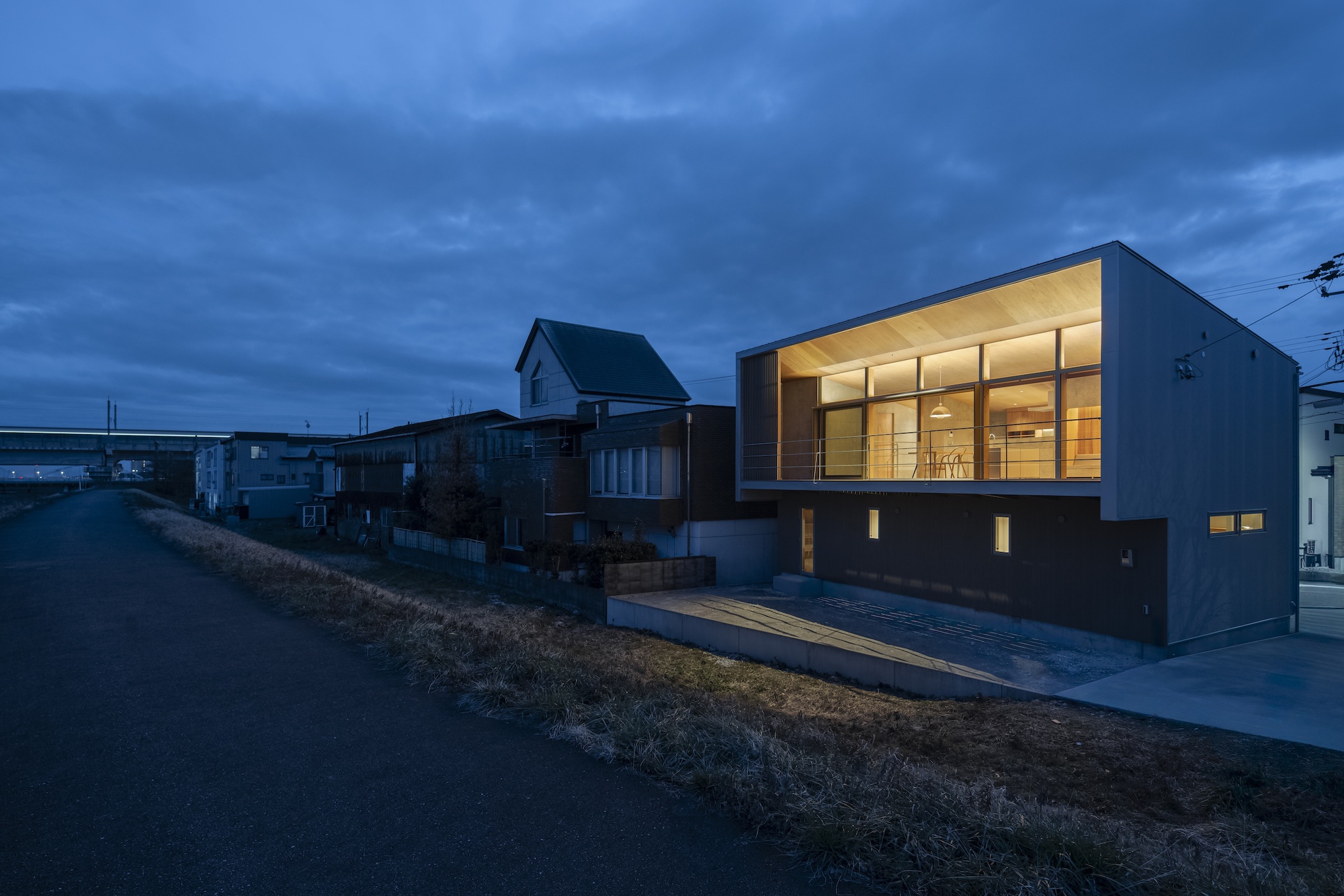 Two-story residence with illuminated glass facade overlooking neighboring houses at dusk