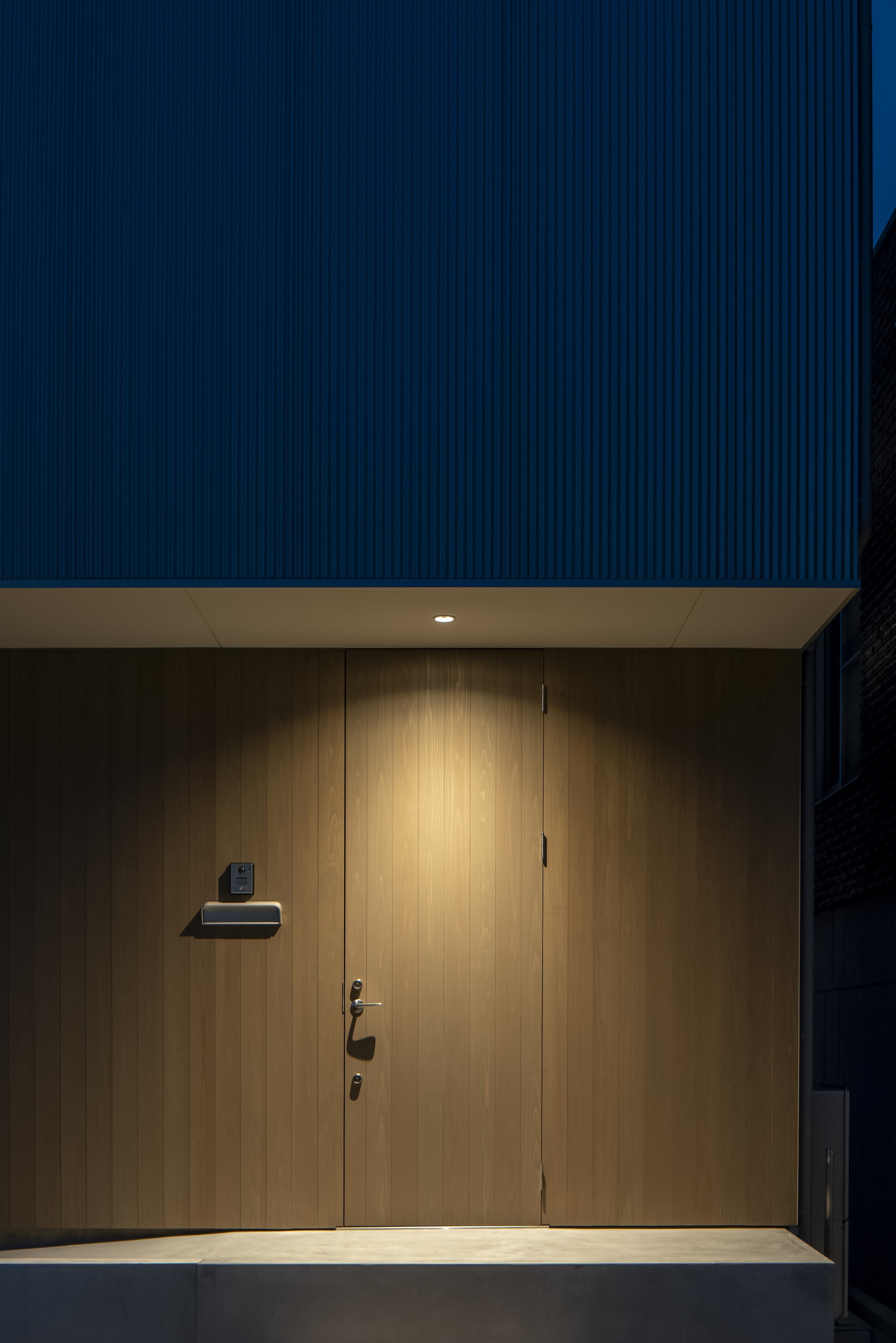 Recessed entry with vertical timber door and mailbox under a dark corrugated metal soffit