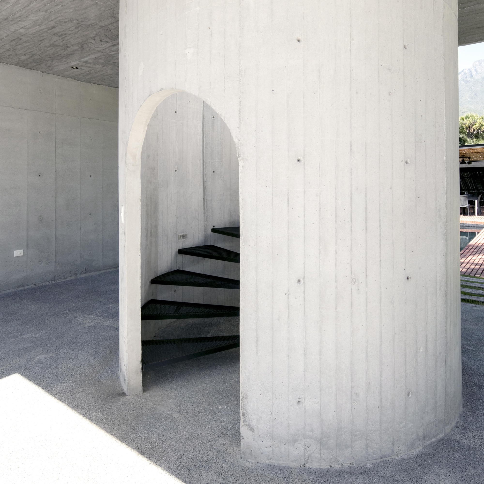 Cylindrical concrete core with arched opening enclosing a black spiral staircase in dappled sunlight