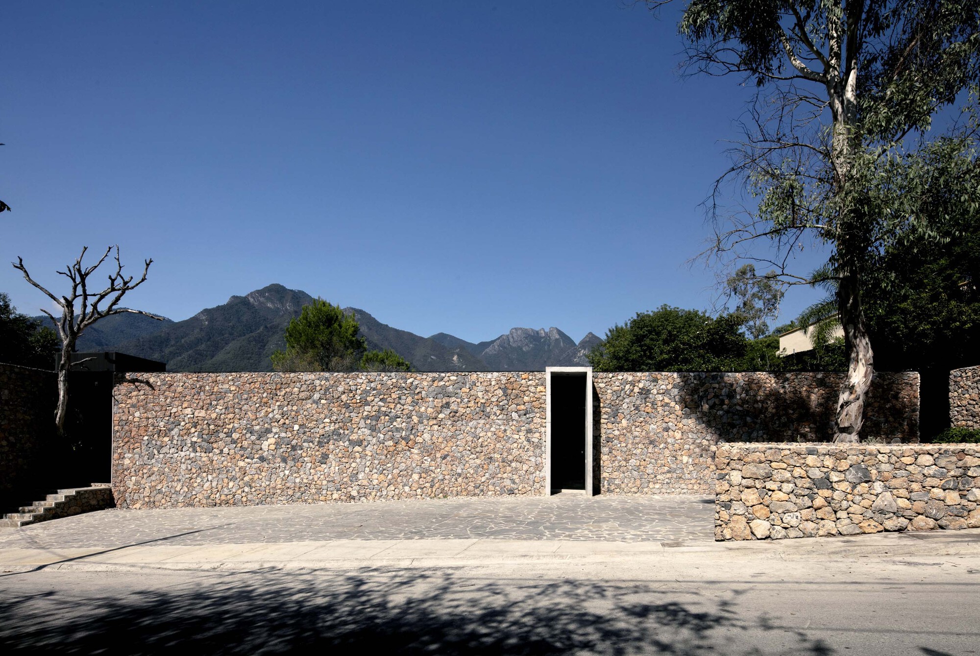 Gabion stone wall with black recessed doorway and mountain range visible in the distance