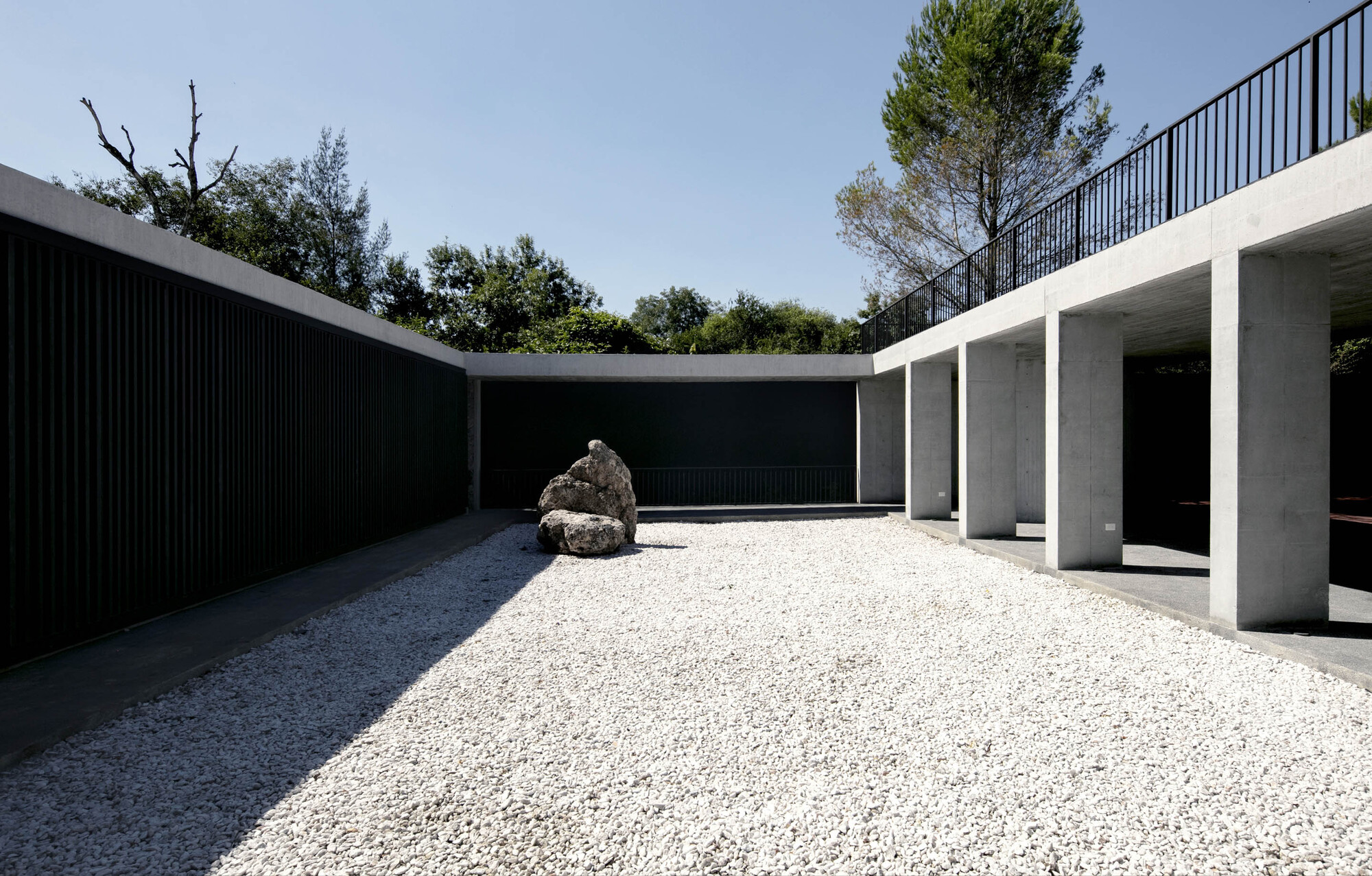 Gravel courtyard with a single boulder between concrete colonnade and black slatted wall