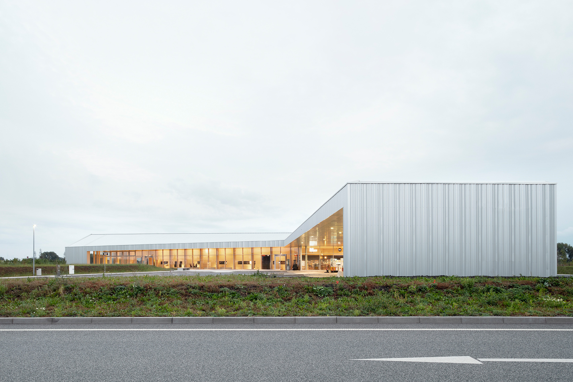 Long metal-clad volume with glazed timber pavilion viewed from the adjacent roadway at dusk