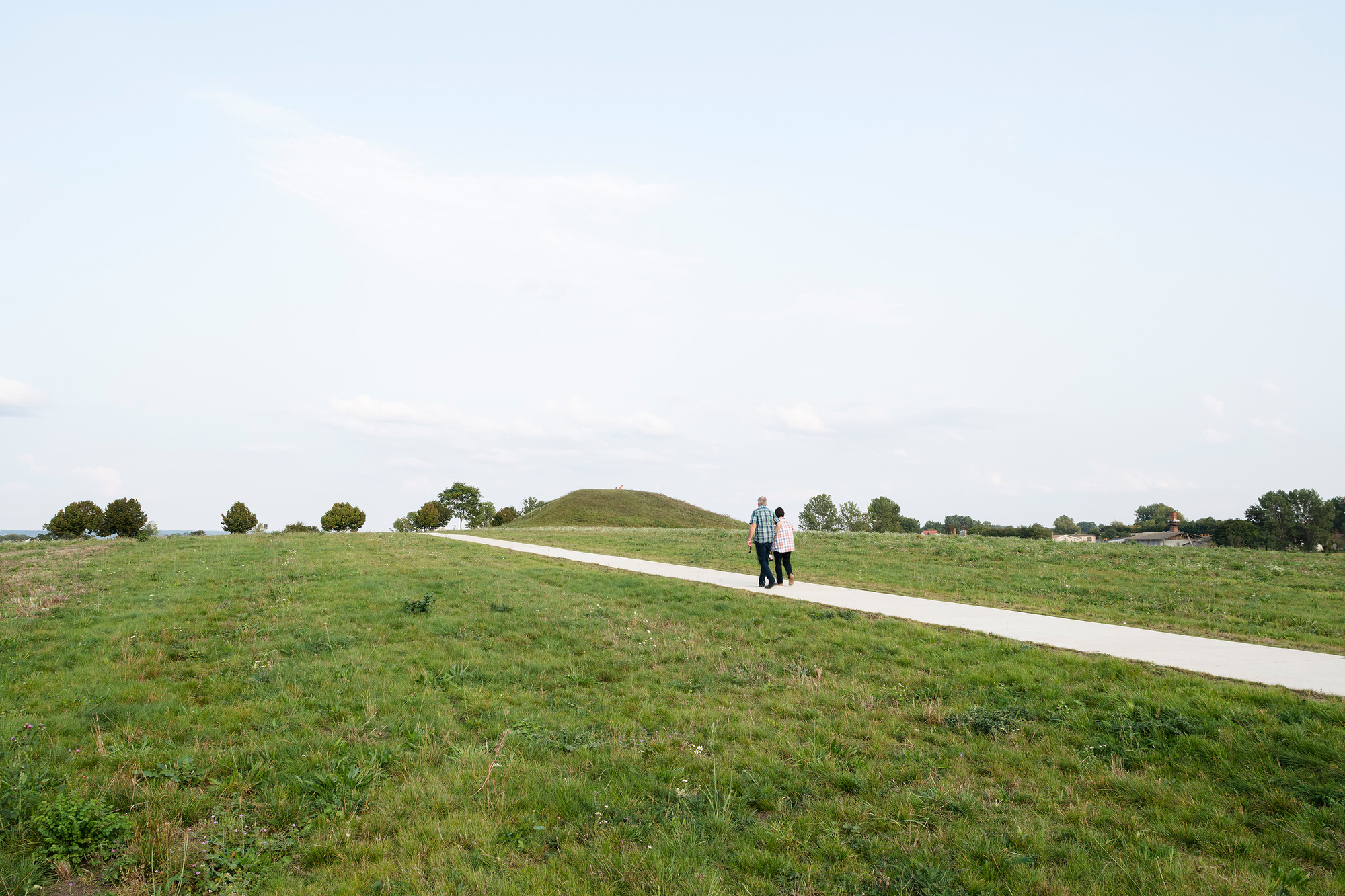 Concrete path crossing a green meadow with two people walking toward a distant mound