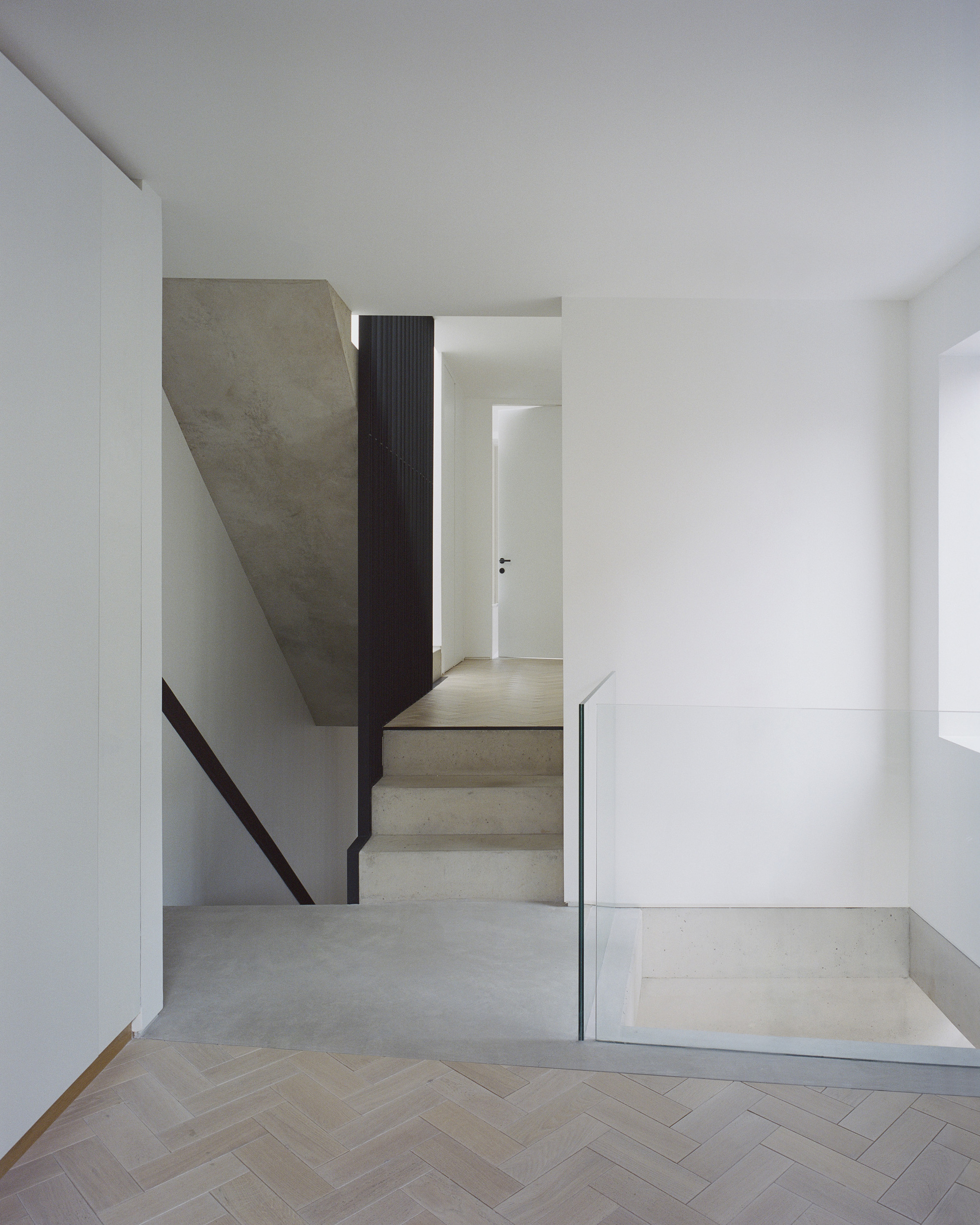 Concrete staircase with glass and metal guardrail descending to herringbone timber floor