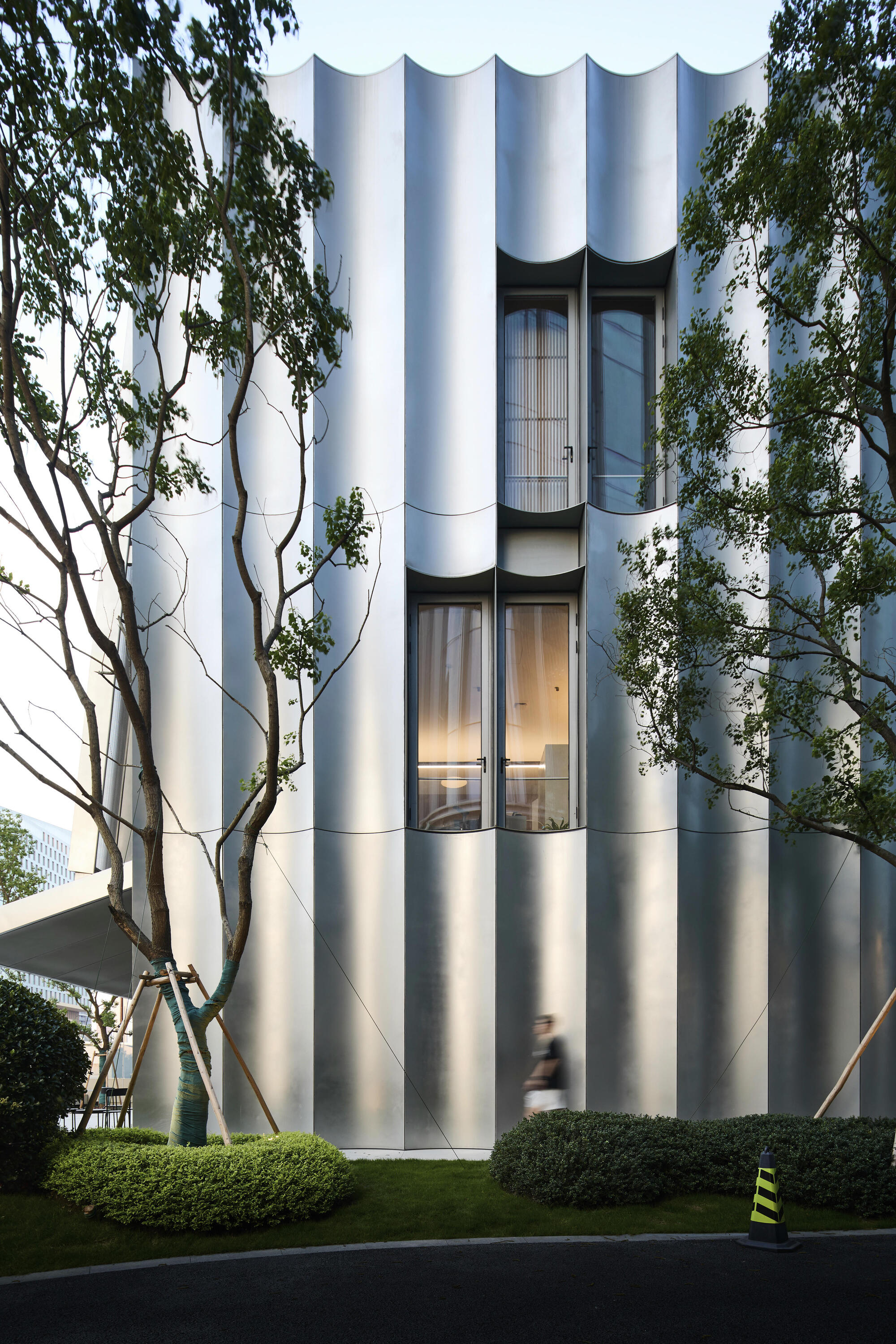 Cylindrical tower with fluted white facade and recessed glazed openings framed by bare trees at dusk