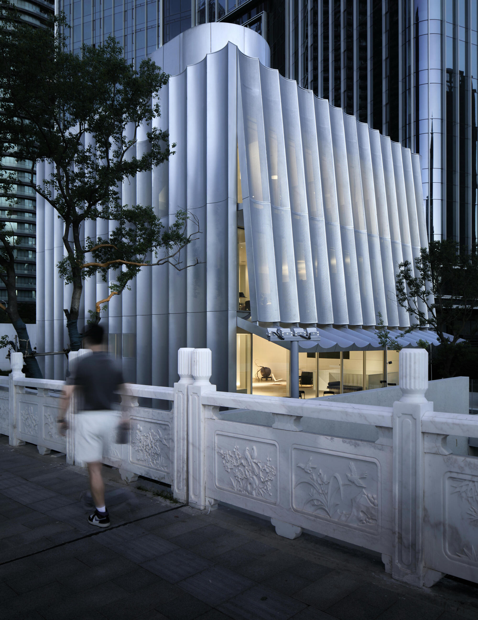 Fluted white volume with illuminated ground floor and pedestrian passing carved stone balustrade at dusk