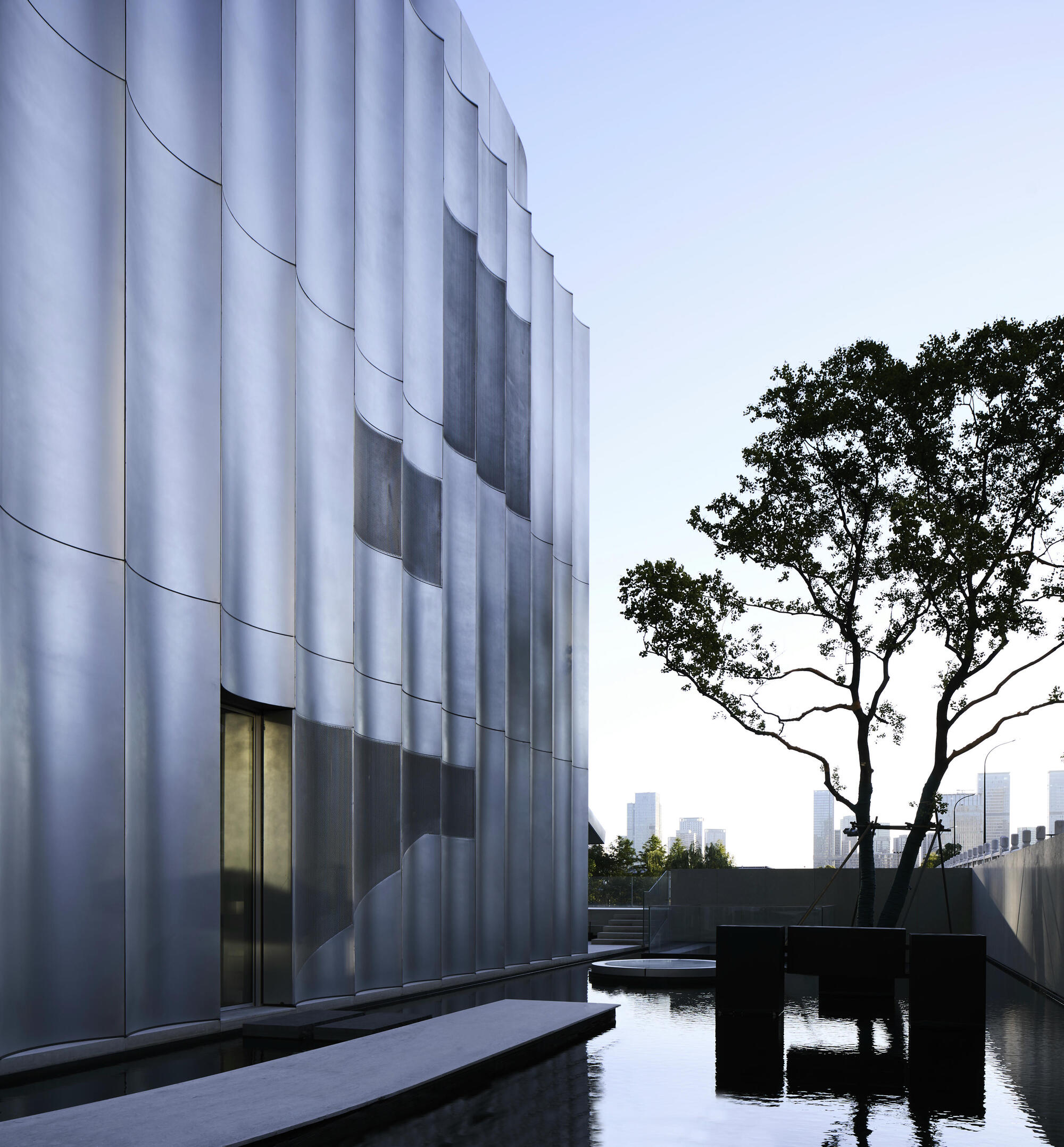 Close-up of white fluted facade with recessed glazing beside a silhouetted tree at twilight