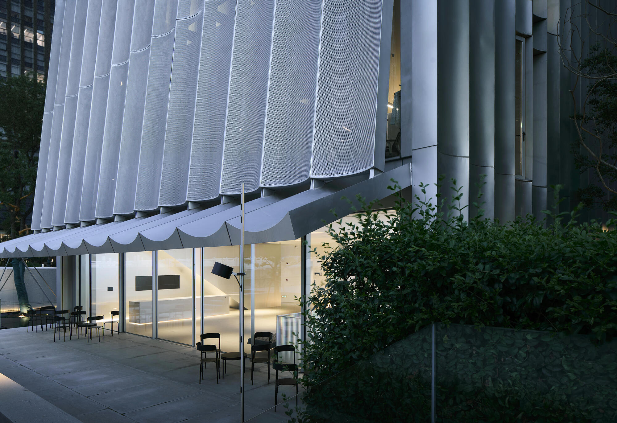 Glass-walled ground floor beneath the scalloped white screen facade at dusk with chairs on the terrace