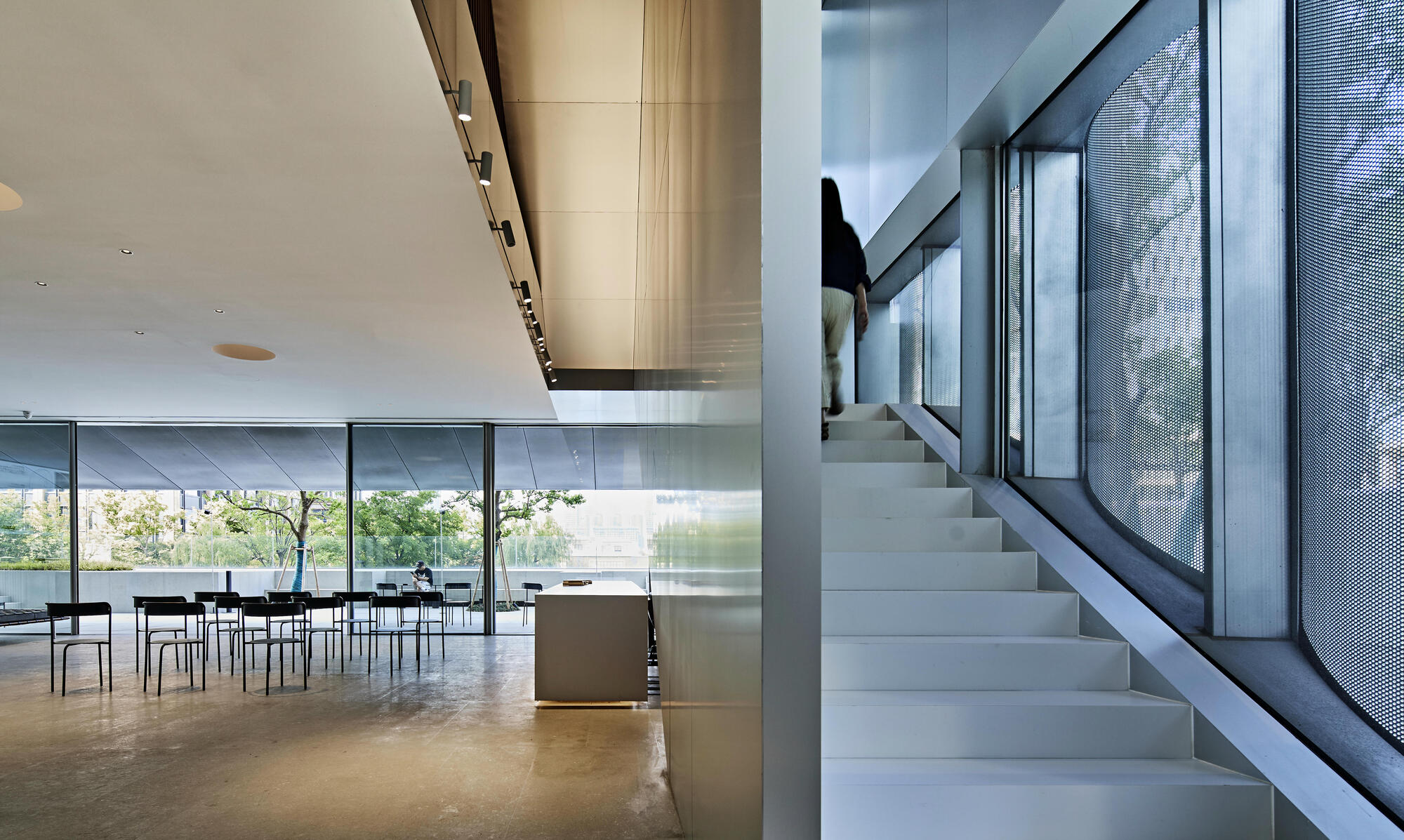 White terrazzo staircase with steel handrail ascending alongside perforated metal screen and glazed courtyard beyond