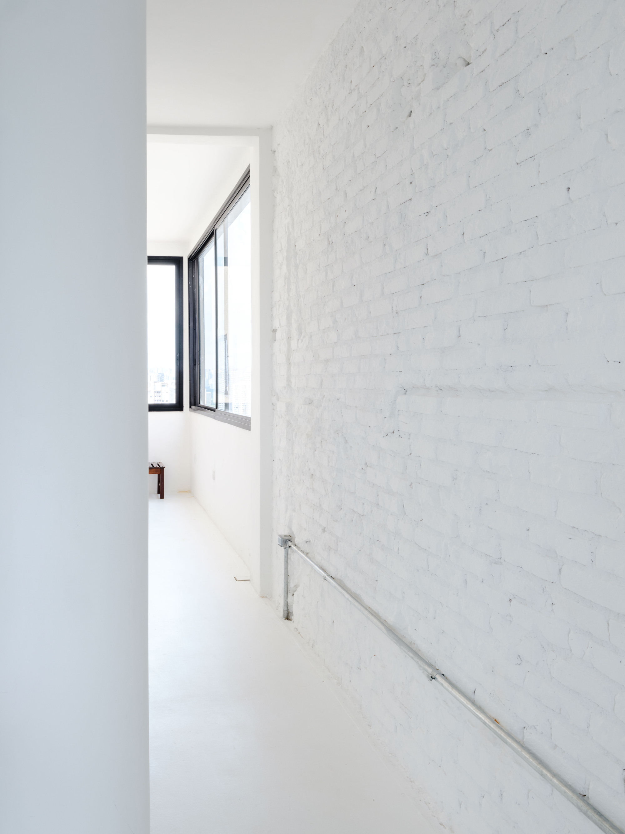 Narrow hallway with white-painted brick wall and stainless steel handrail leading toward a black-framed window