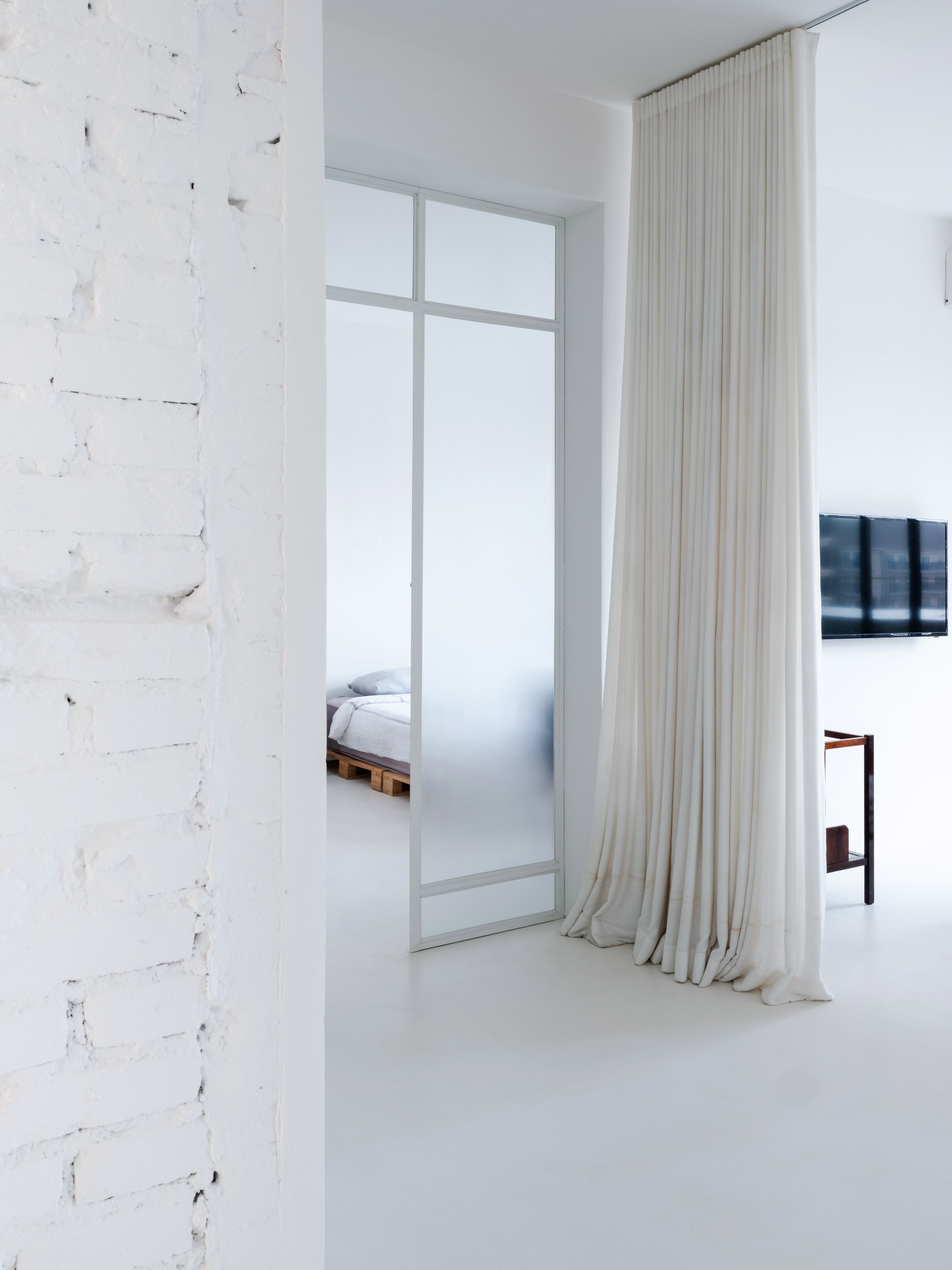 White interior with distressed brick wall, floor-to-ceiling sheer curtains and glimpse of bedroom beyond frosted glass partition