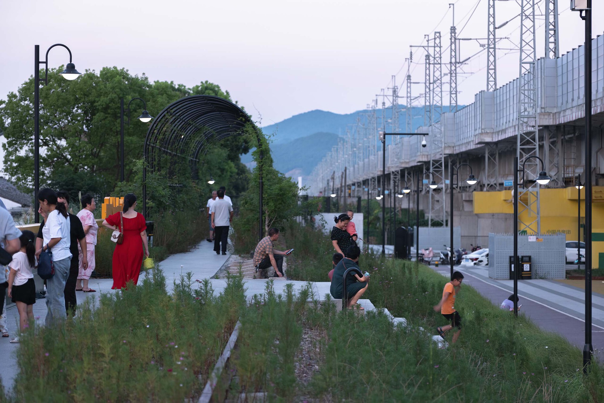 Linear pedestrian walkway with planted beds and visitors gathering near the arched trellis at twilight