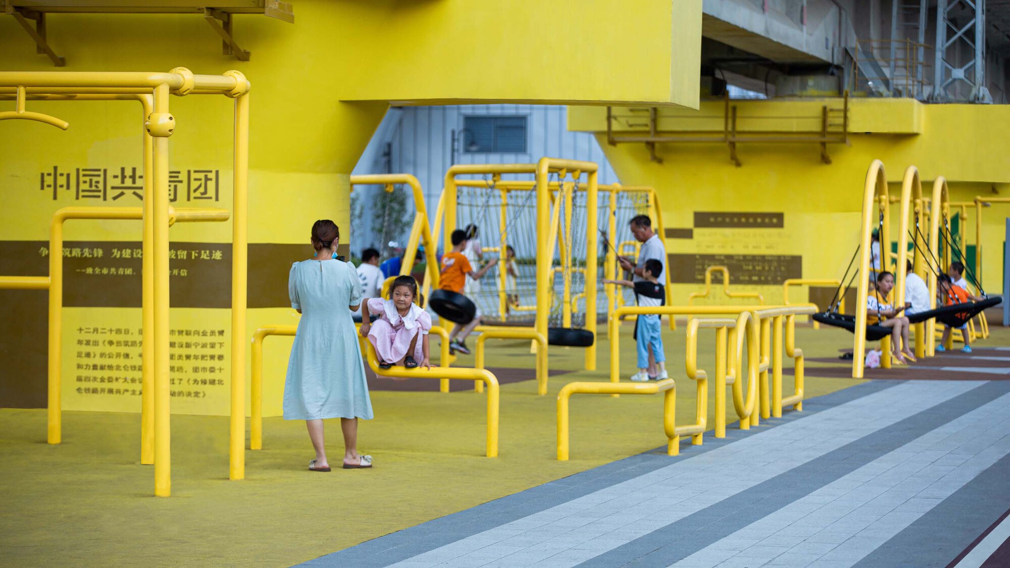 Yellow tubular playground structures with children and families playing beneath the concrete viaduct