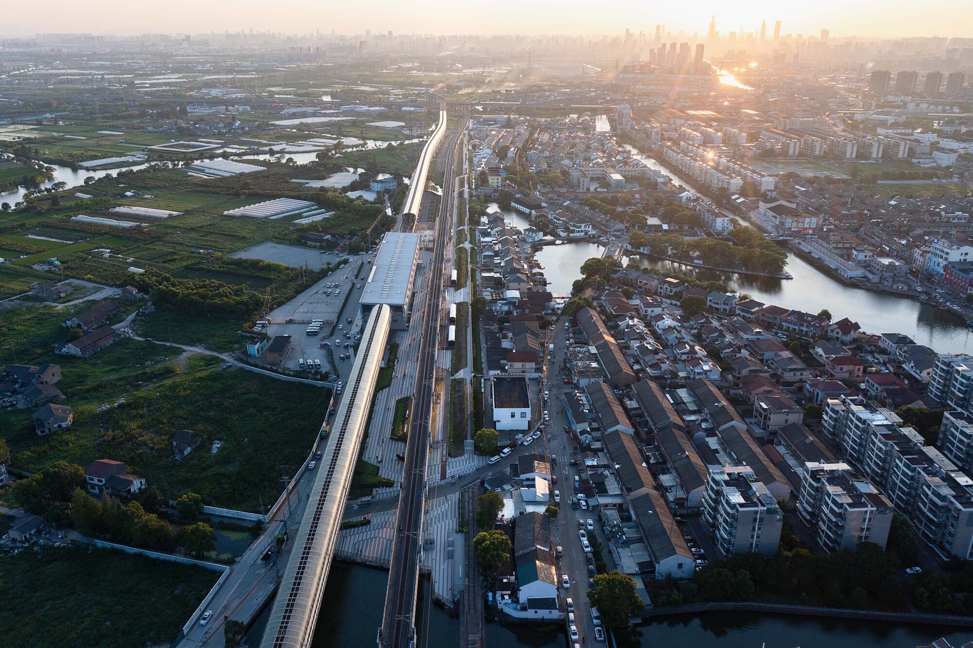 Drone view of the rail corridor and station canopy threading through wetlands and housing at sunset
