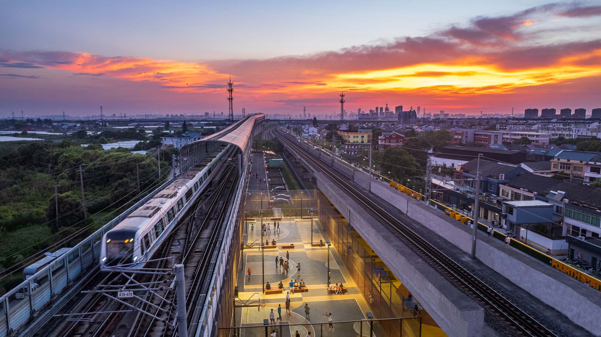 Elevated rail station with sunken glass-walled platform lit from within beneath converging tracks at sunset