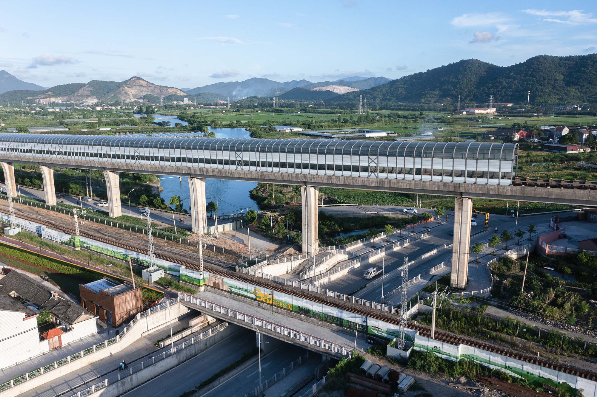 Multi-level rail viaduct with arched tubular canopy crossing roadways and green fields below distant mountains