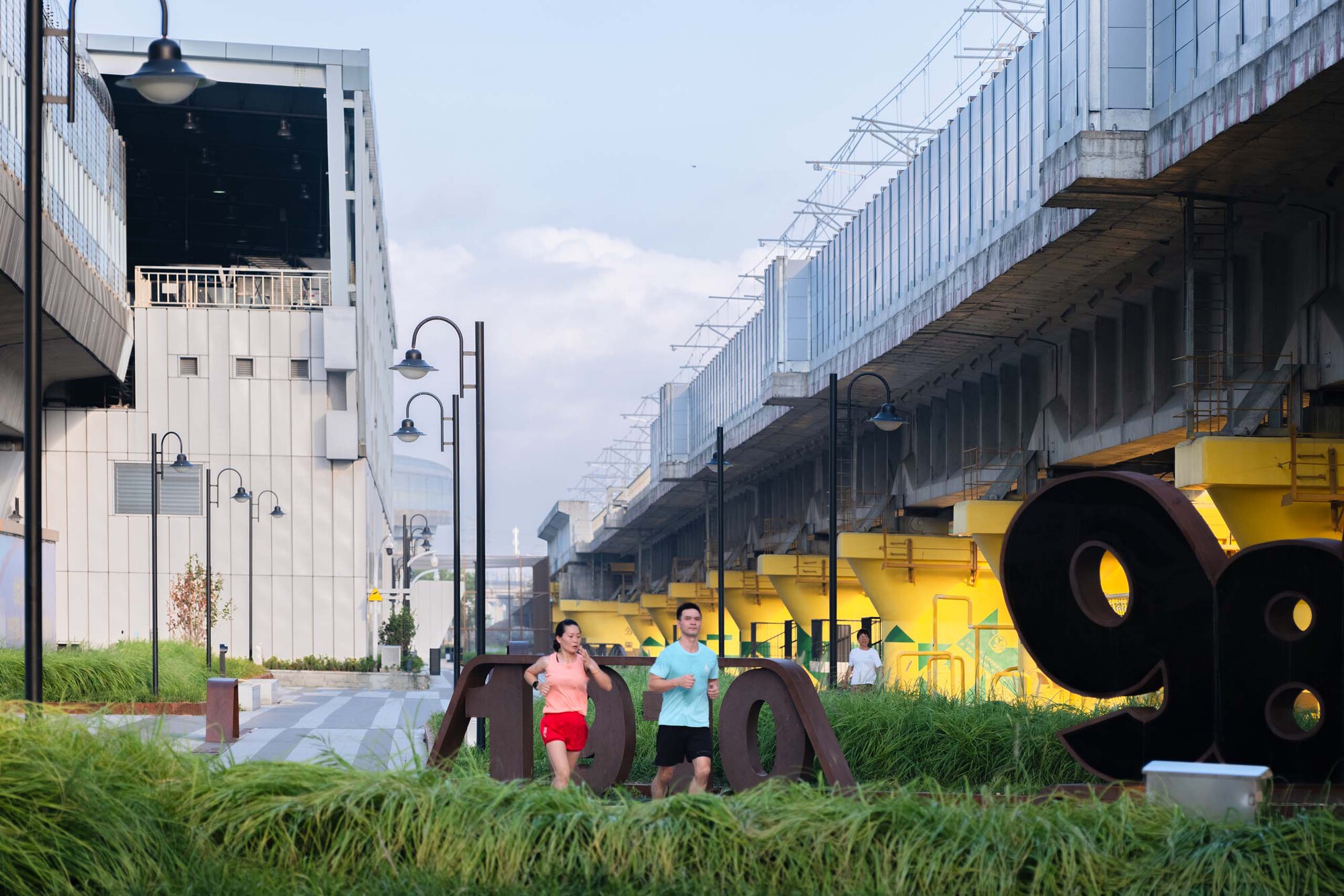 Ground-level plaza with yellow sculptural elements and joggers passing beneath elevated rail infrastructure at dusk