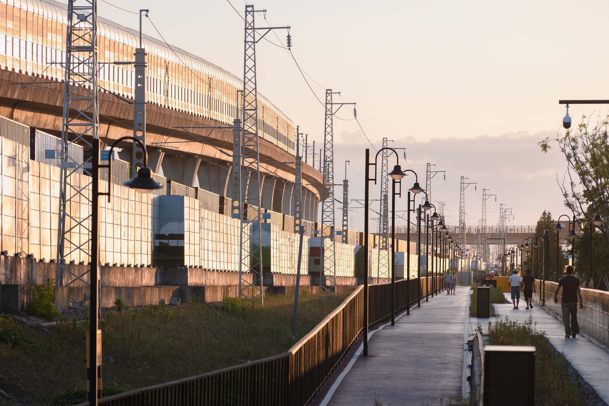 Pedestrian walkway along corrugated metal station wall with streetlights and people strolling at golden hour