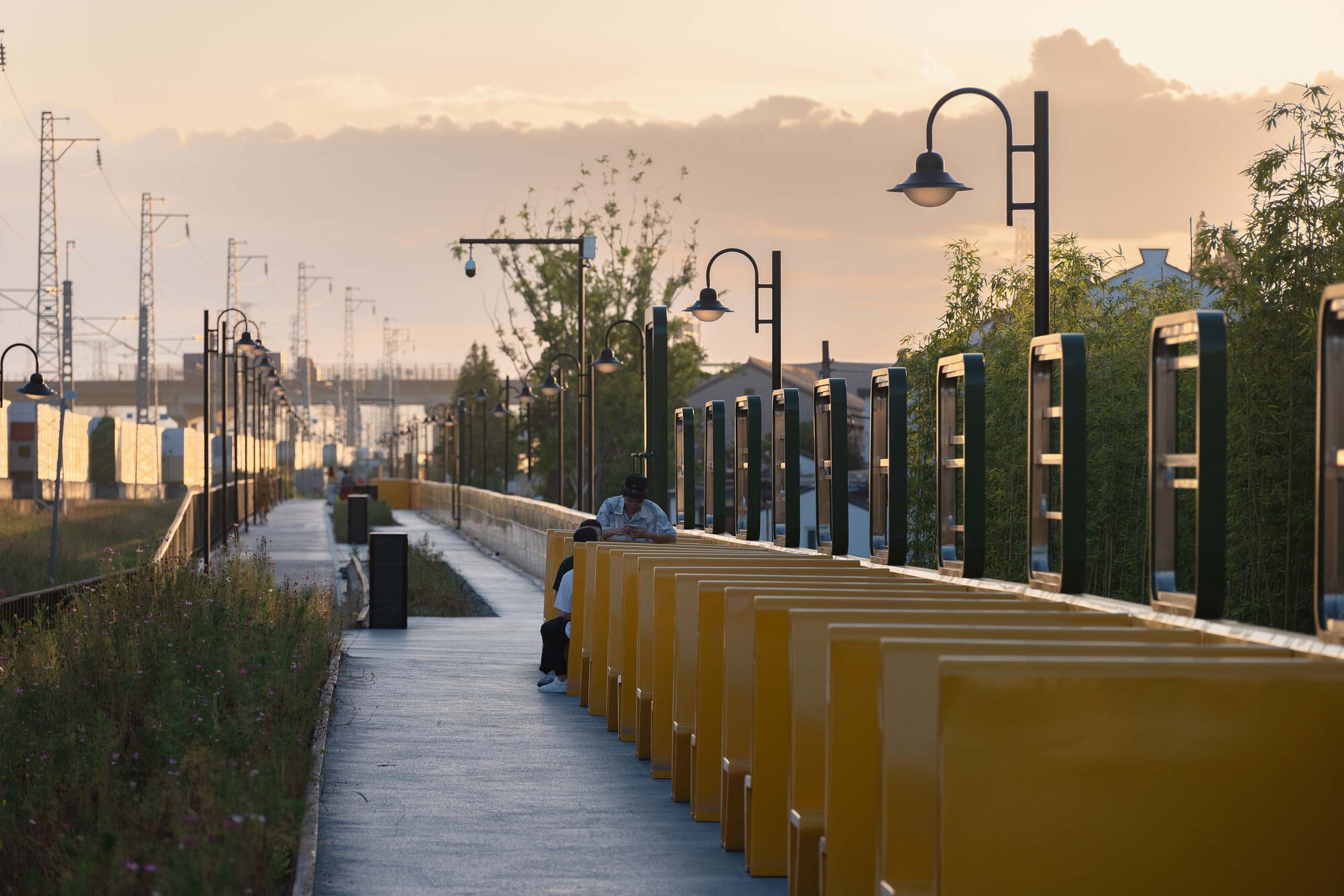 Linear promenade with repeating yellow benches and metal viewing frames along railway edge at golden hour