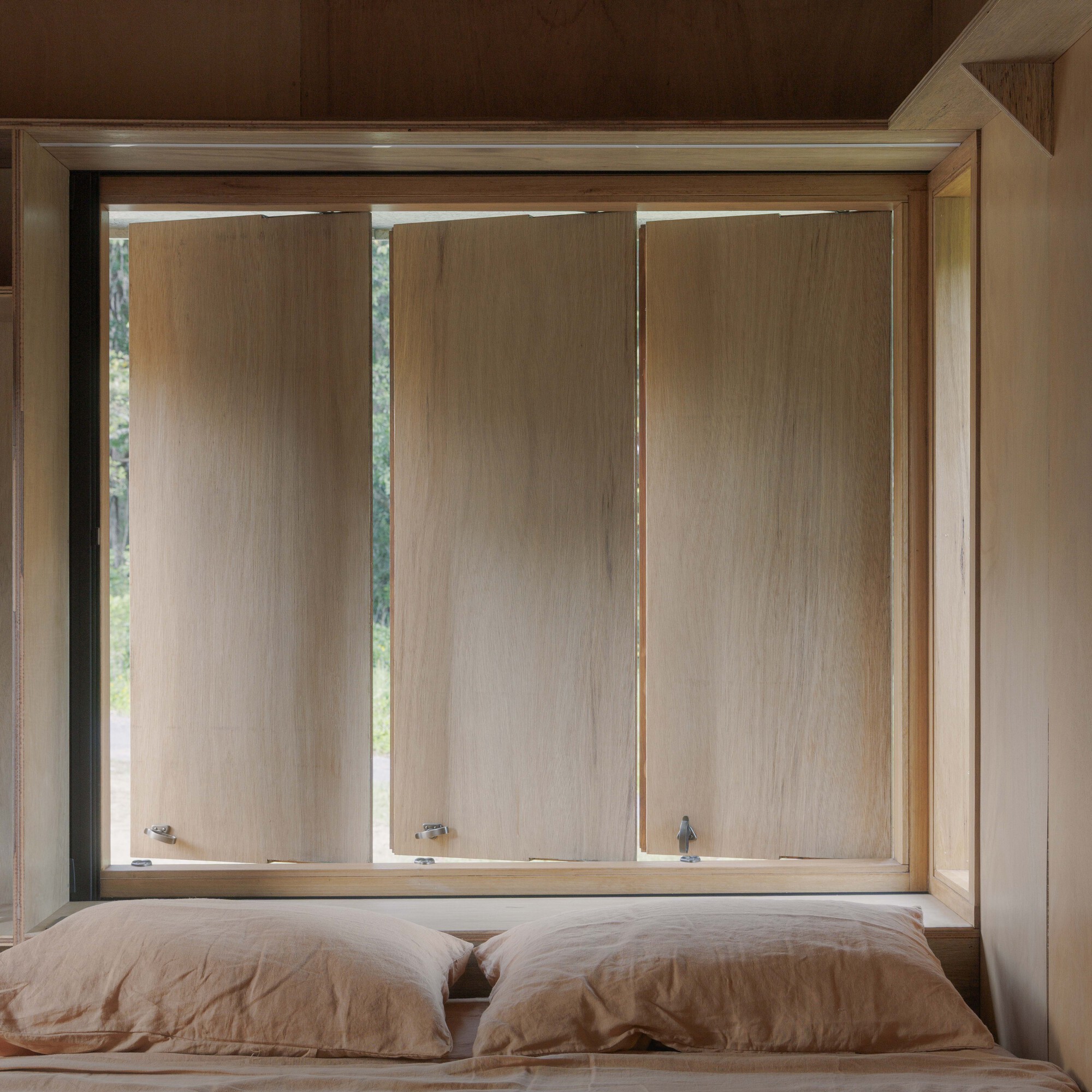 Bedroom window with three vertical timber panels framed in plywood above a linen-covered bed