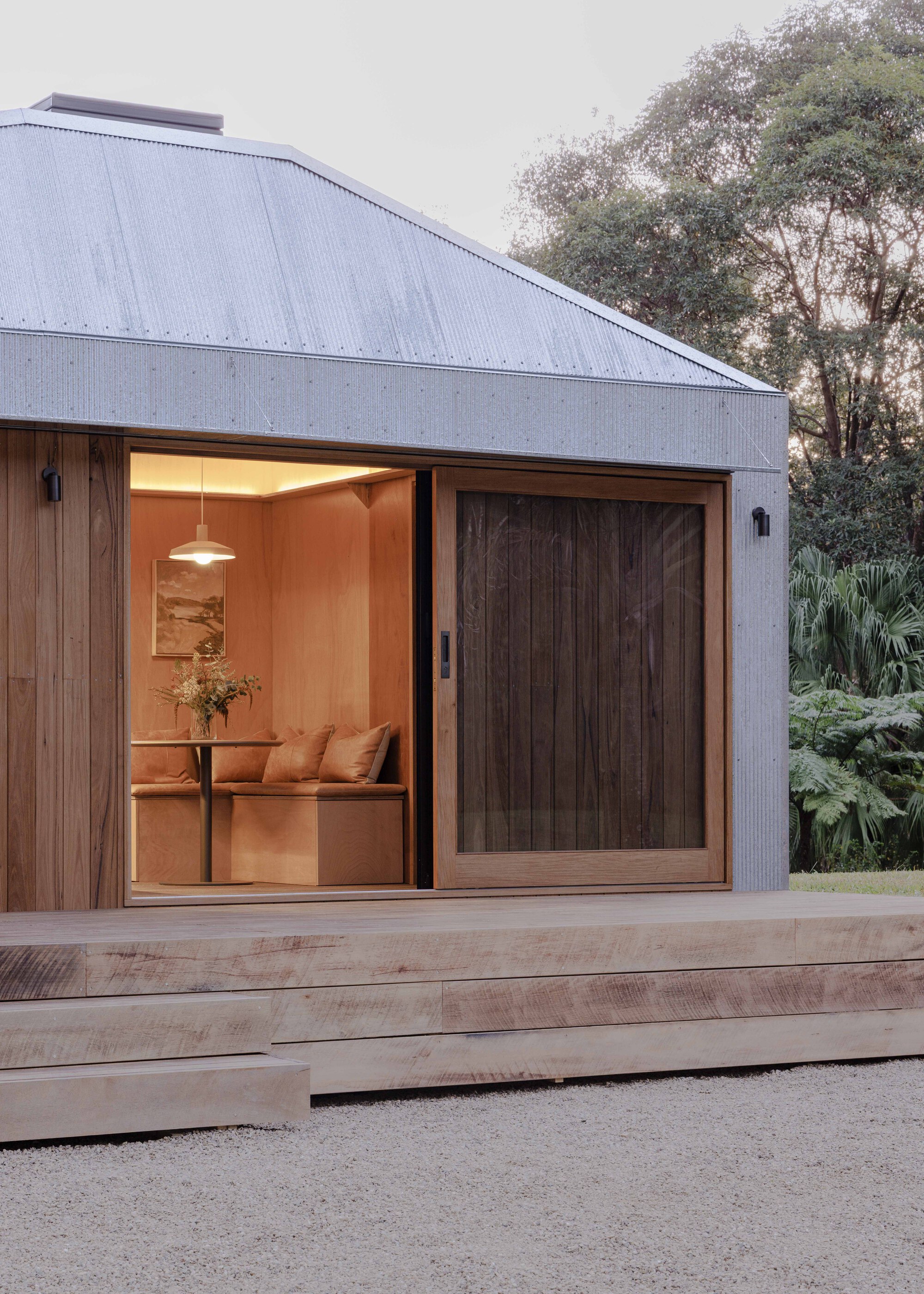 Corner of the pavilion at dusk showing sliding timber door and glowing interior under metal roof