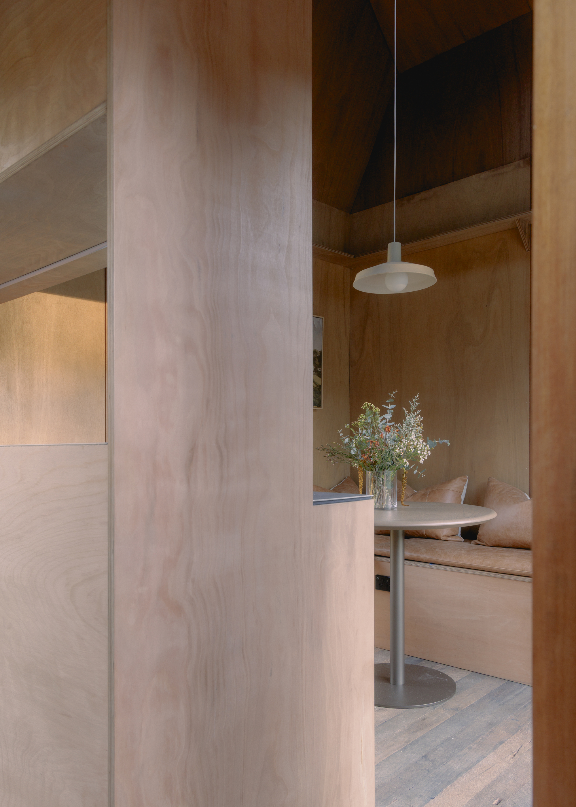 View through plywood-clad corridor toward the dining area with pendant light and vase of wildflowers