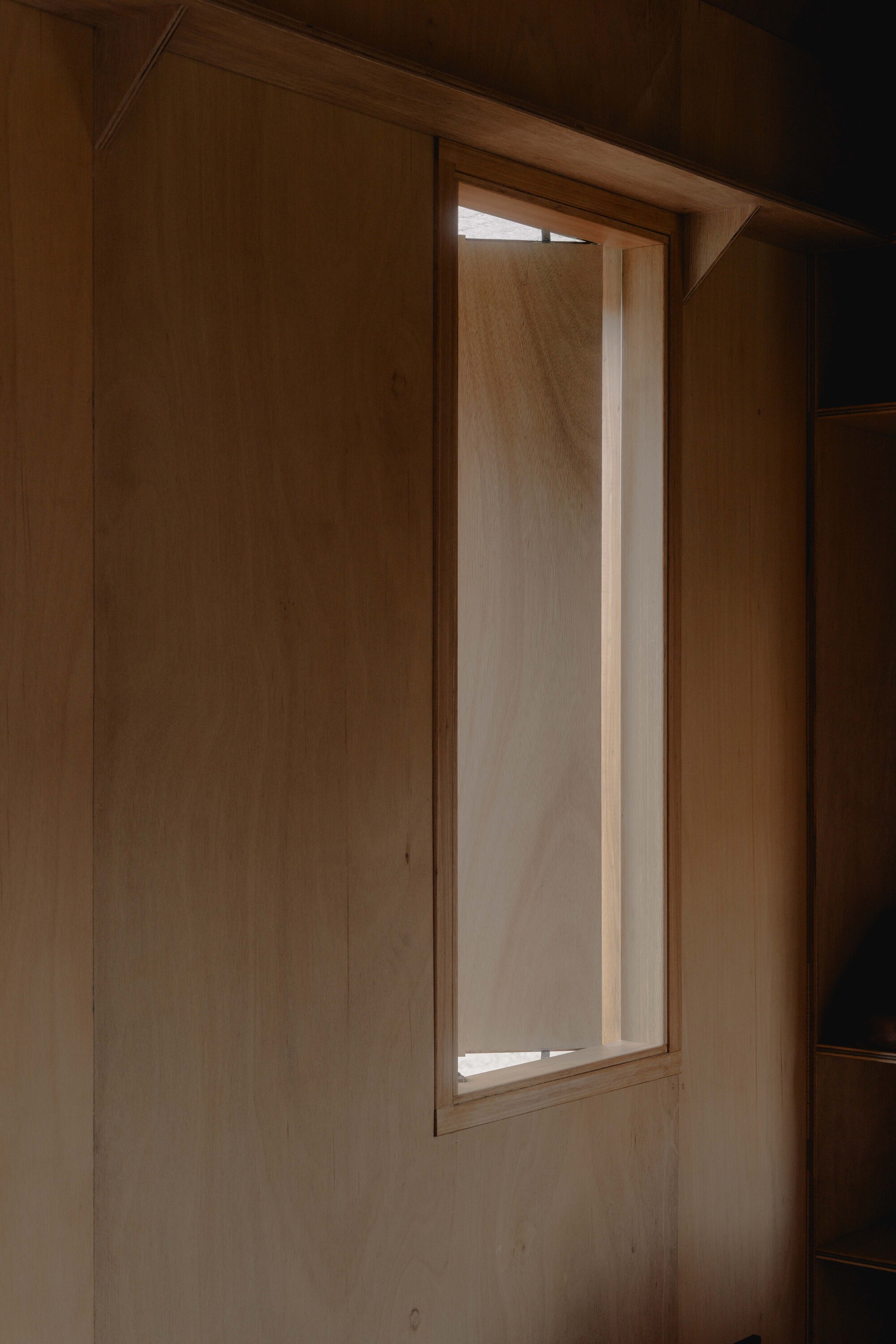 Tall narrow window set into plywood wall softly illuminated at dusk