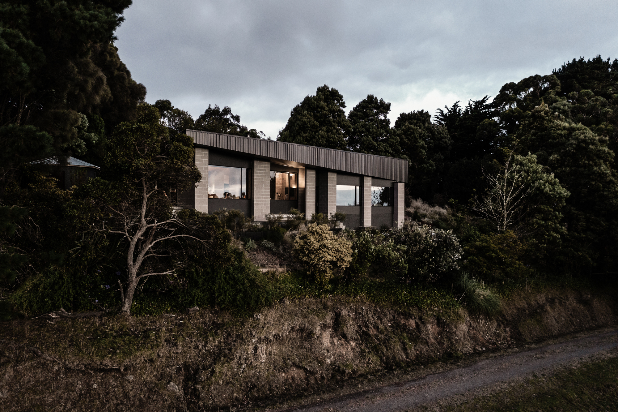 Elevated house with timber and concrete volumes perched above a sloped hillside under overcast skies