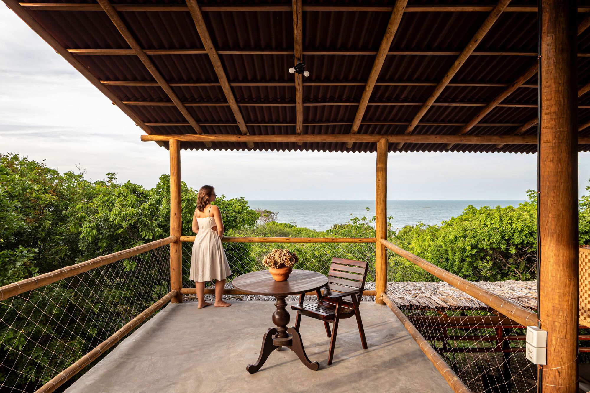 Upper terrace with rope railing where a person stands overlooking the ocean through coastal vegetation