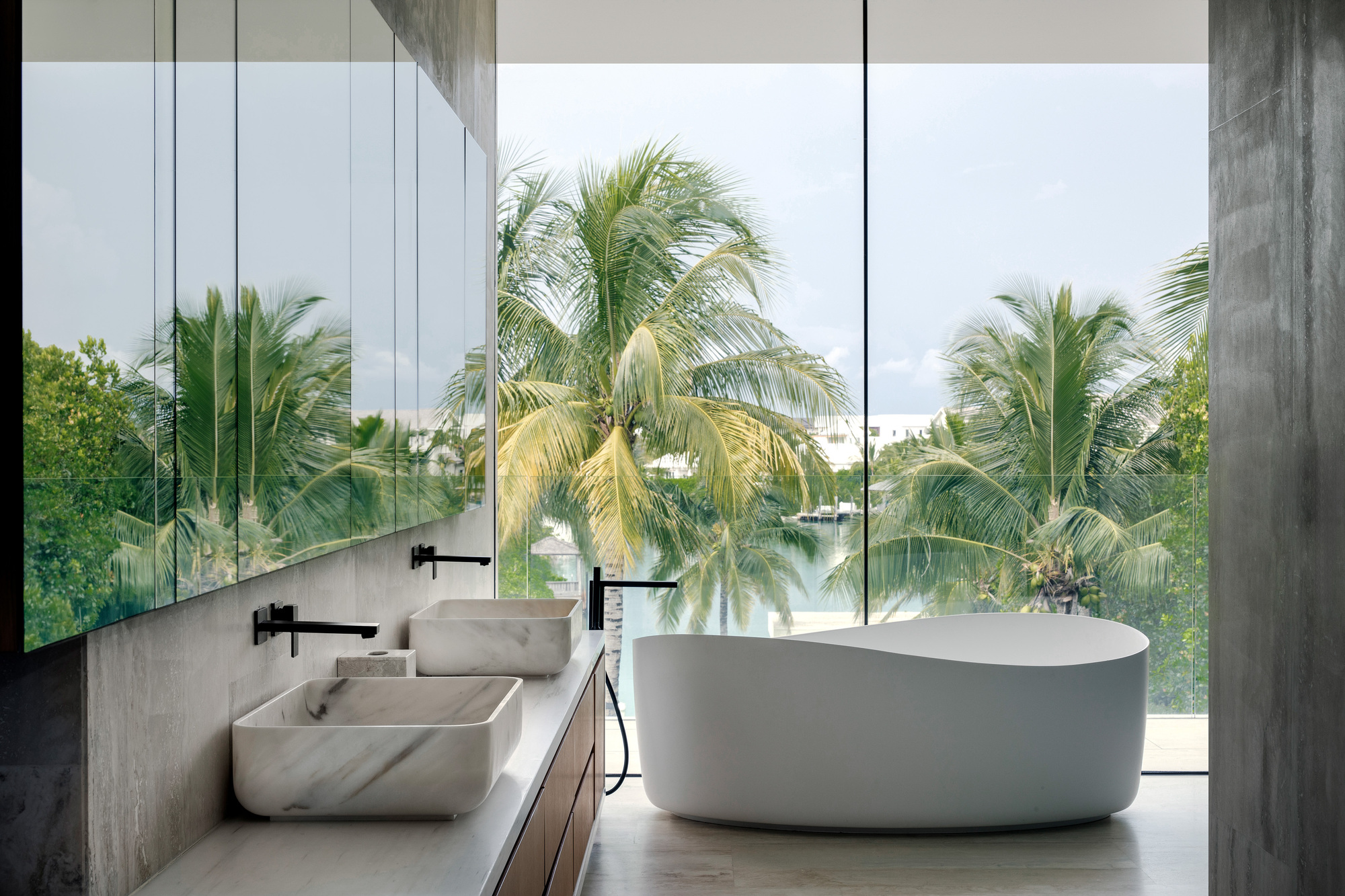 Bathroom interior with freestanding tub and dual stone basins opening to palm trees through floor-to-ceiling glass