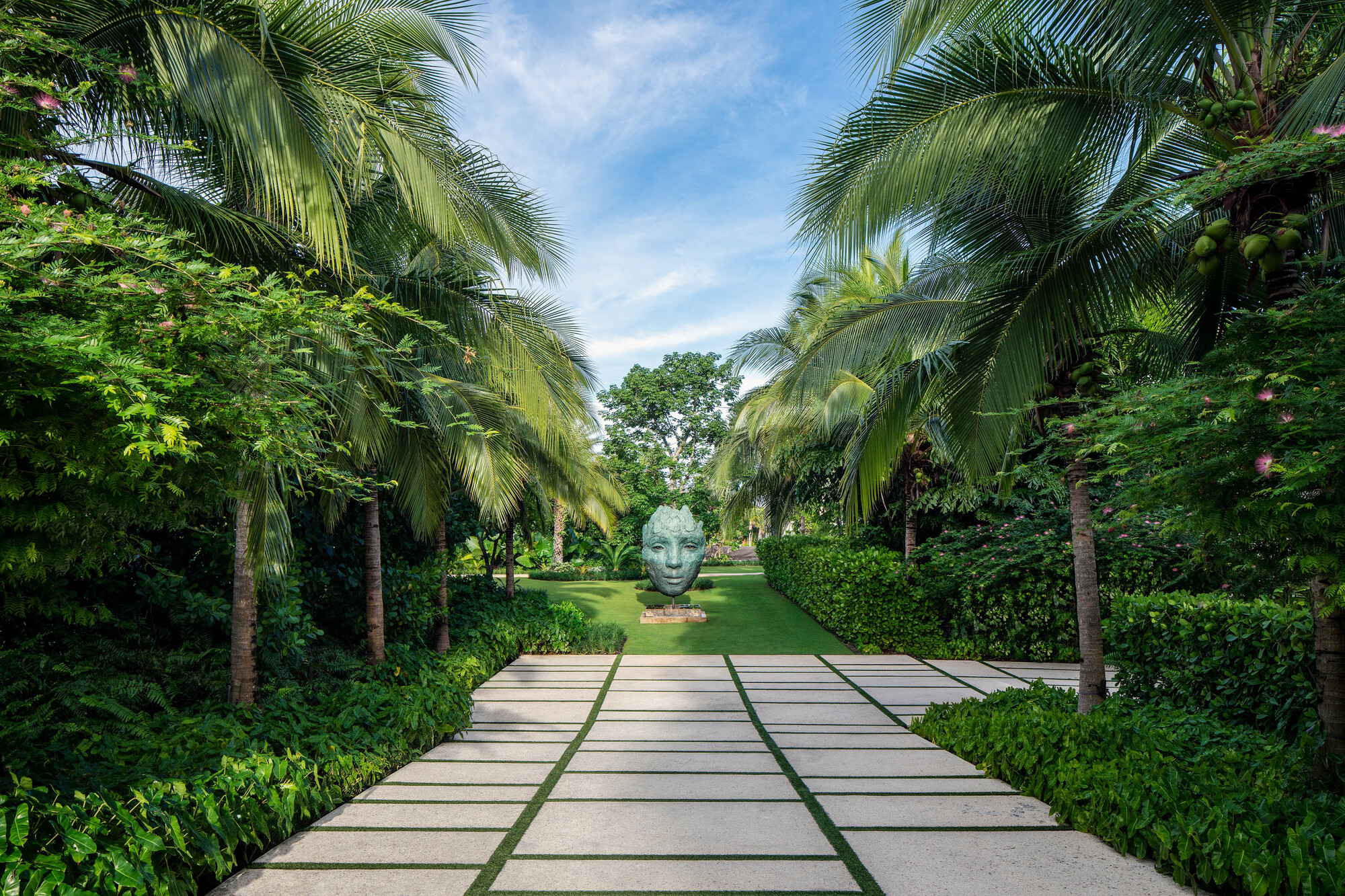 Paved garden path with grass joints leading to a sculptural head beneath palm canopy and blue sky